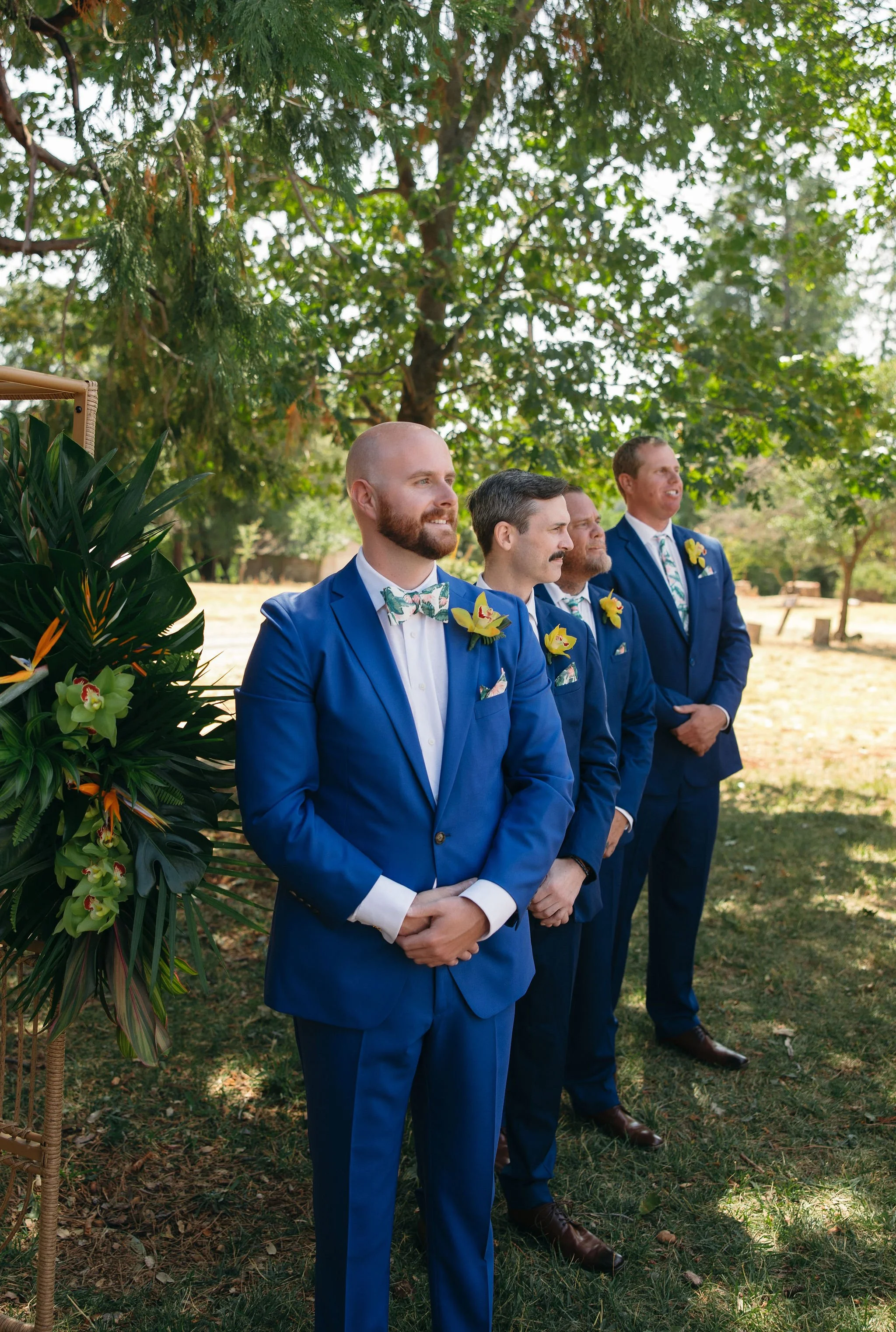 Four men in blue suits stand outdoors during a wedding ceremony, with a large green tree and floral decorations nearby.