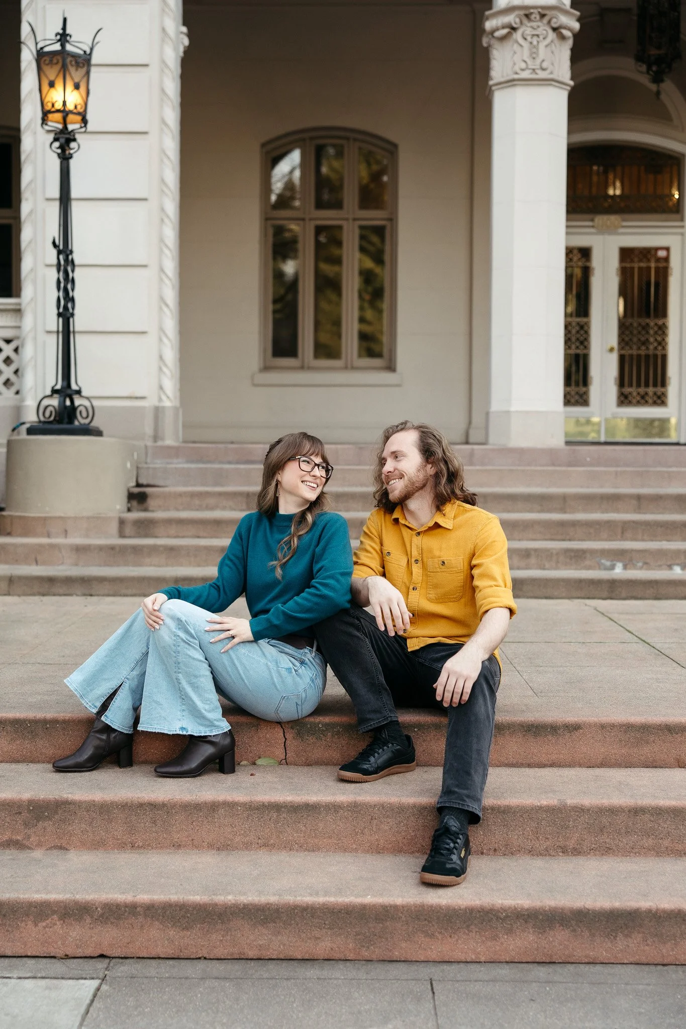 A young woman and man sitting on the steps of a building, smiling and looking at each other. The woman is wearing glasses, a blue sweater, and jeans with black heeled boots. The man is wearing a mustard yellow shirt and black pants, with black shoes.