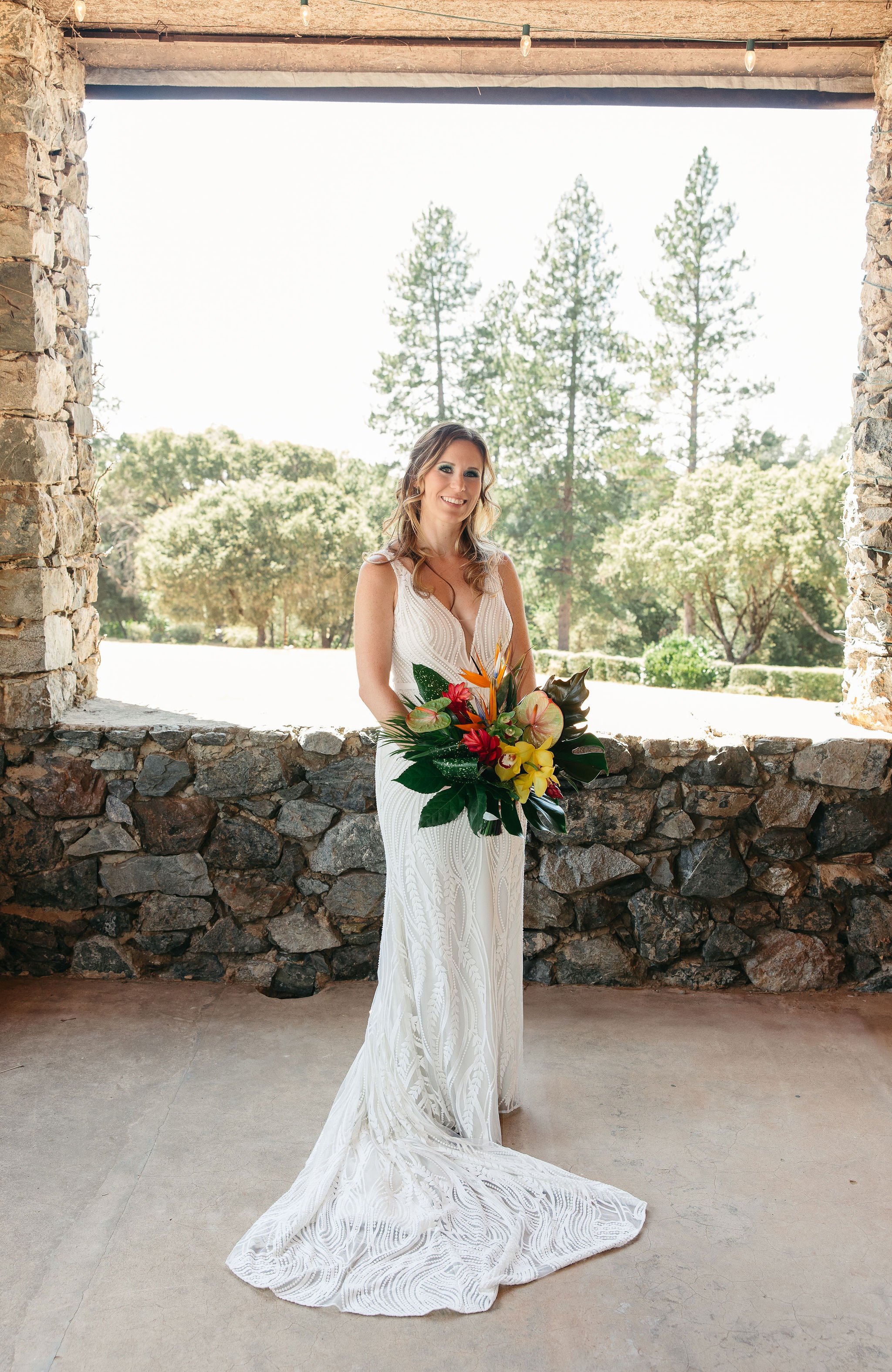 A bride in a white gown with intricate patterns, holding a colorful bouquet of flowers, standing in front of a stone wall opening with trees and clear sky in the background.