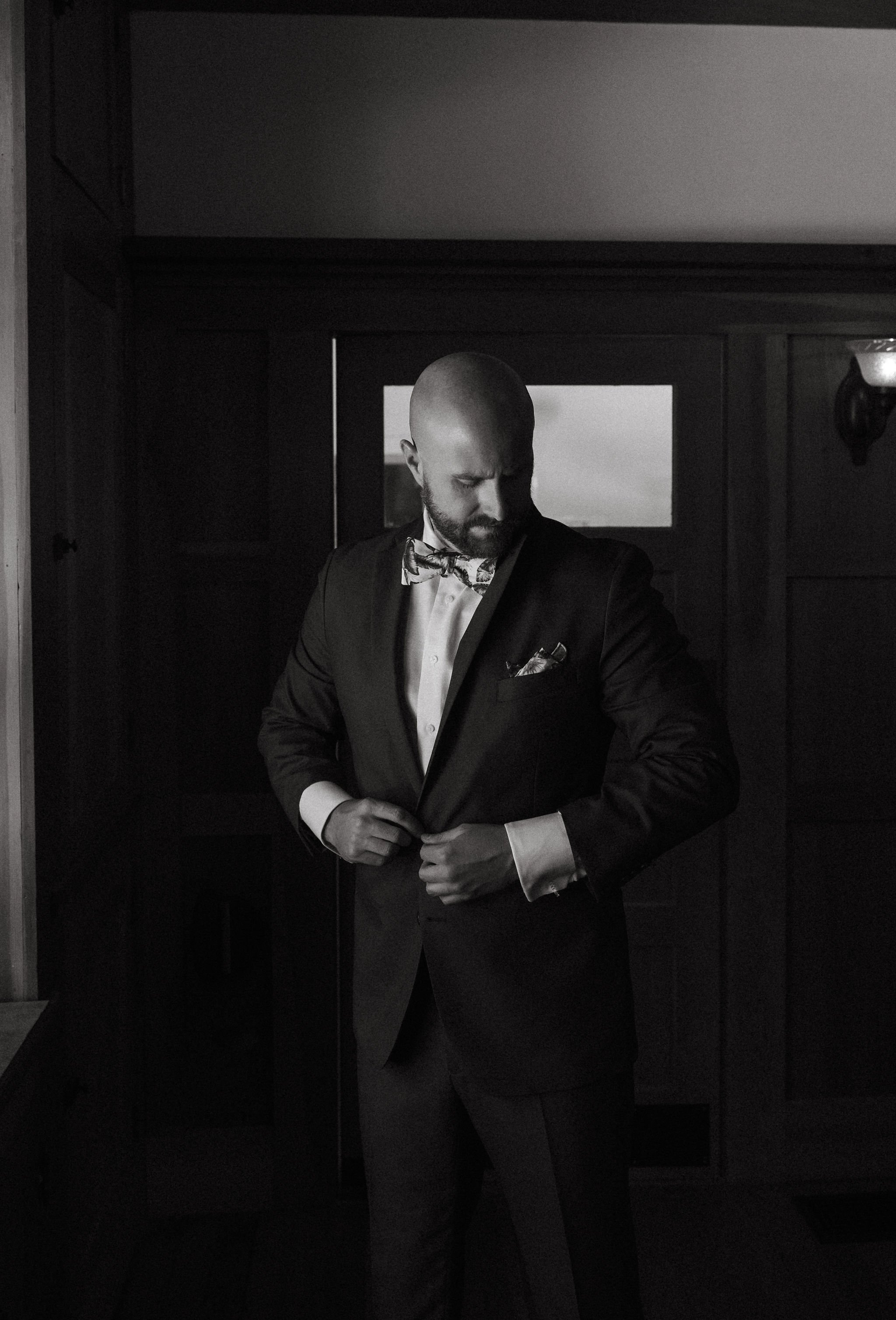 A man in a tuxedo with a bowtie adjusts his jacket in a dimly lit room.