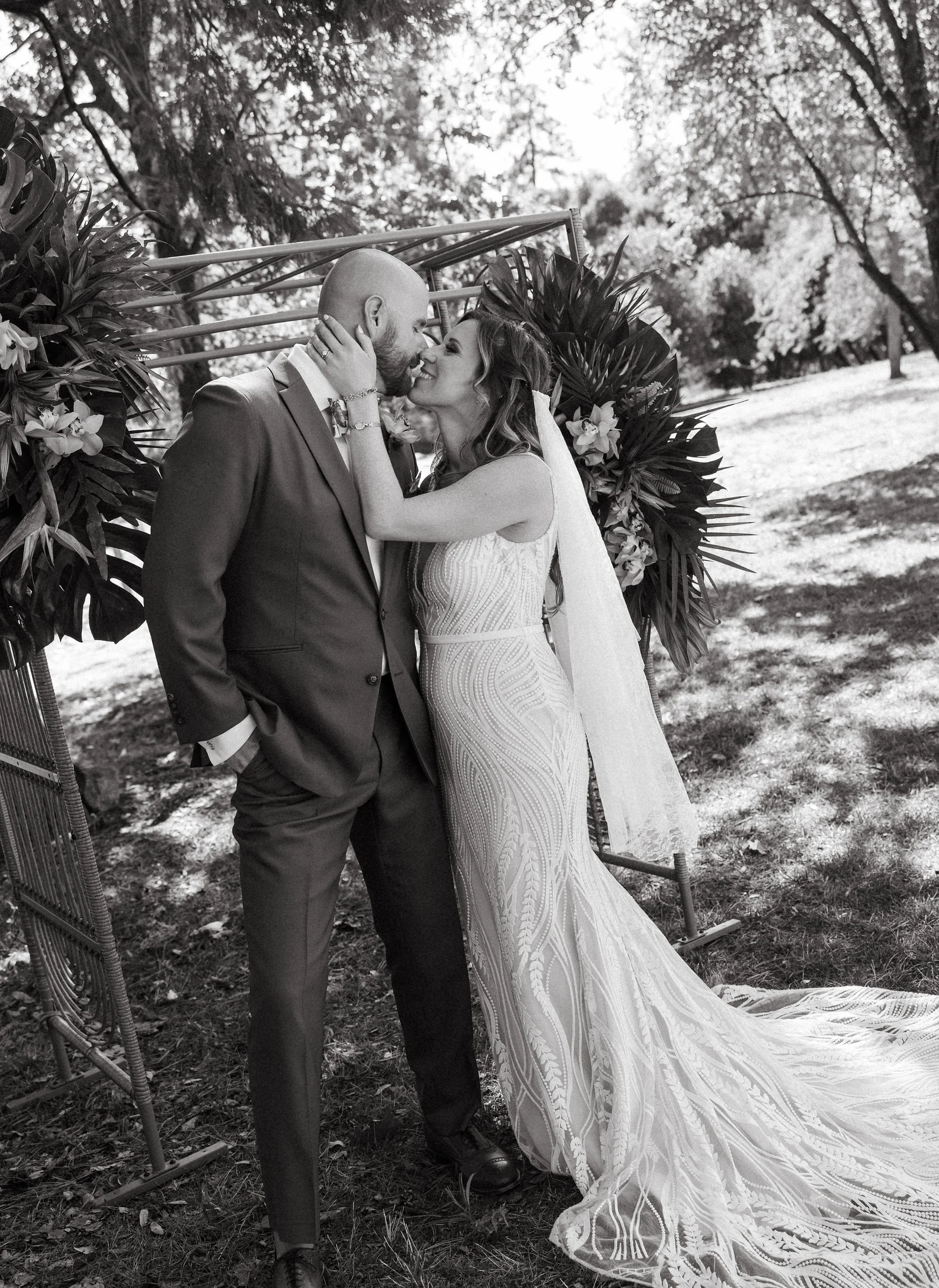 A black-and-white photograph of a bride and groom sharing a kiss outdoors. The bride is wearing a lacy wedding dress with a long train and the groom is in a suit. They are surrounded by trees and decorated with floral arrangements.