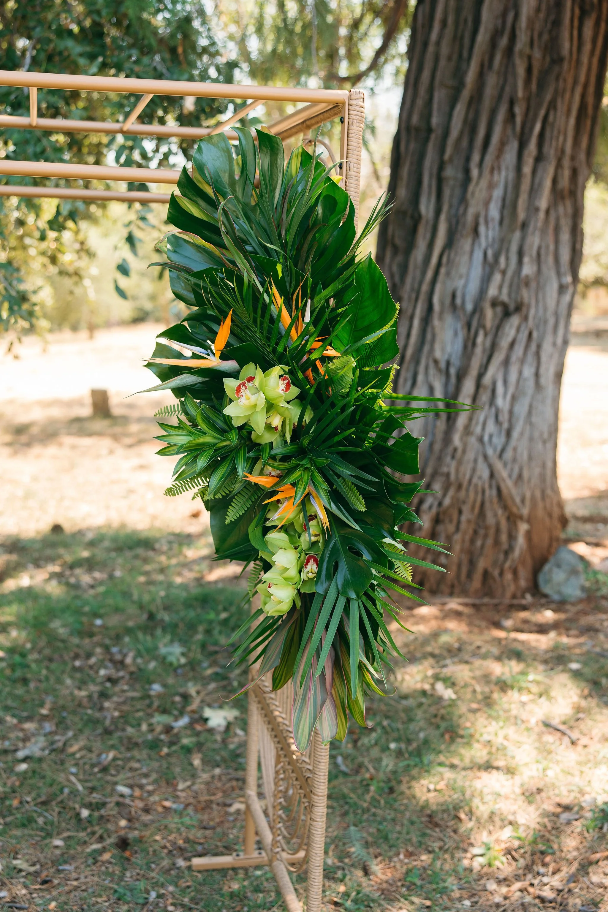 A large floral arrangement with green tropical leaves and white orchids, set outdoors next to a tree and a bamboo stand.