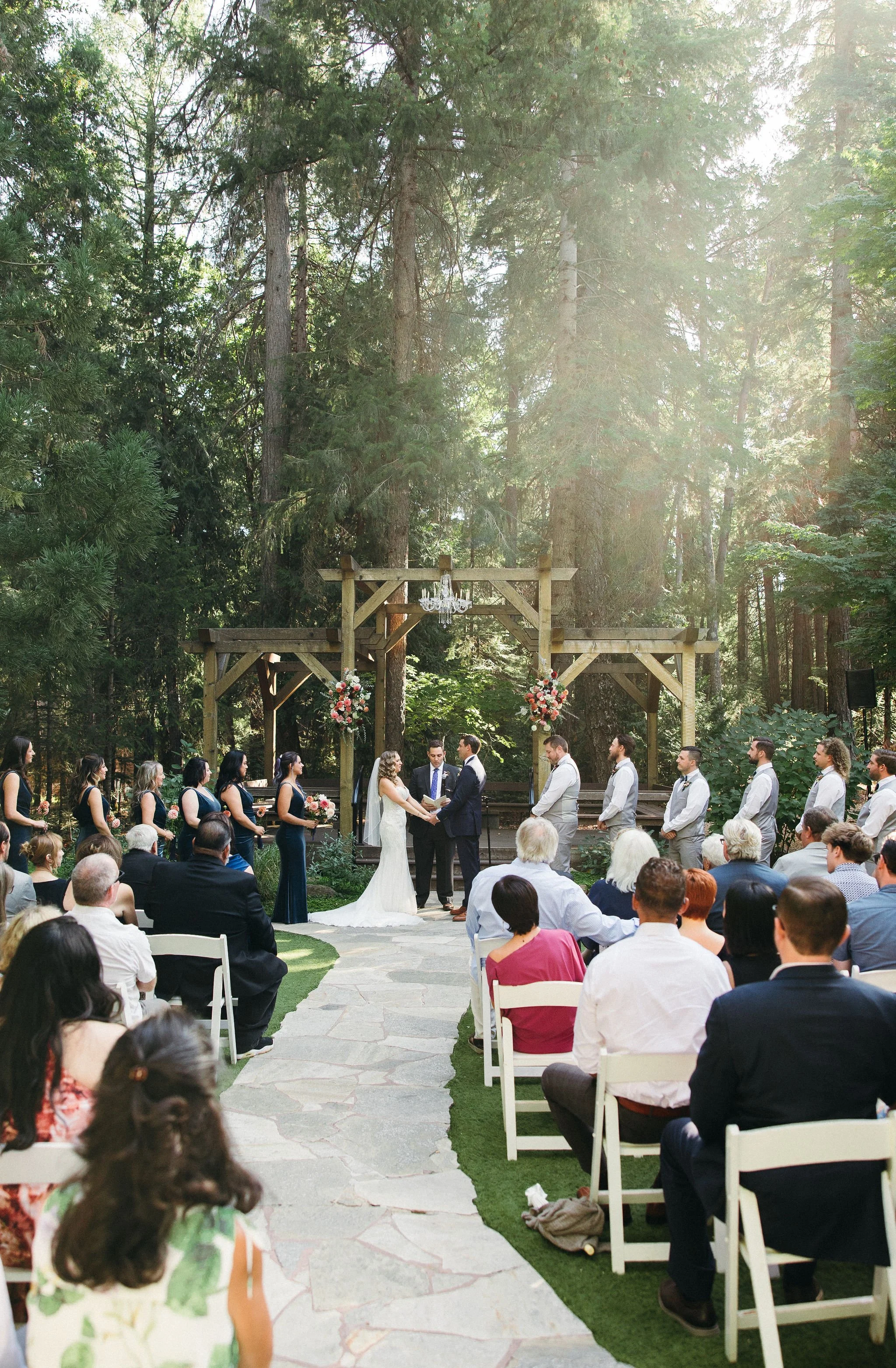 An outdoor wedding ceremony taking place in a forest surrounded by tall trees. The bride and groom are standing under a wooden arch decorated with pink and white flowers, exchanging vows. Guests are seated on white chairs on either side of a stone pa