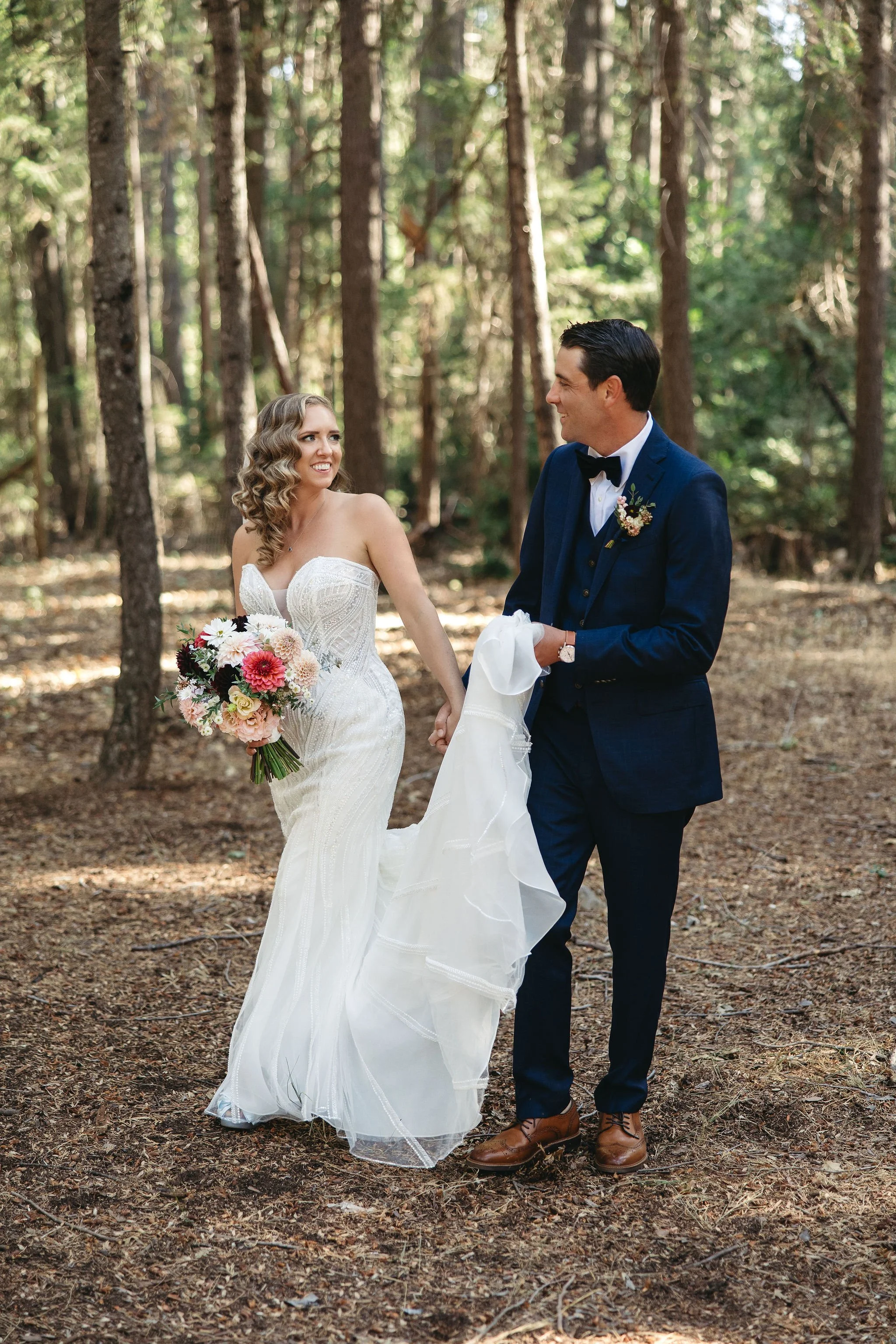 A bride and groom holding hands and smiling at each other in a wooded forest, with sunlight filtering through the trees. The bride is in a strapless white wedding gown holding a bouquet of pink, white, and red flowers. The groom is in a navy blue tux