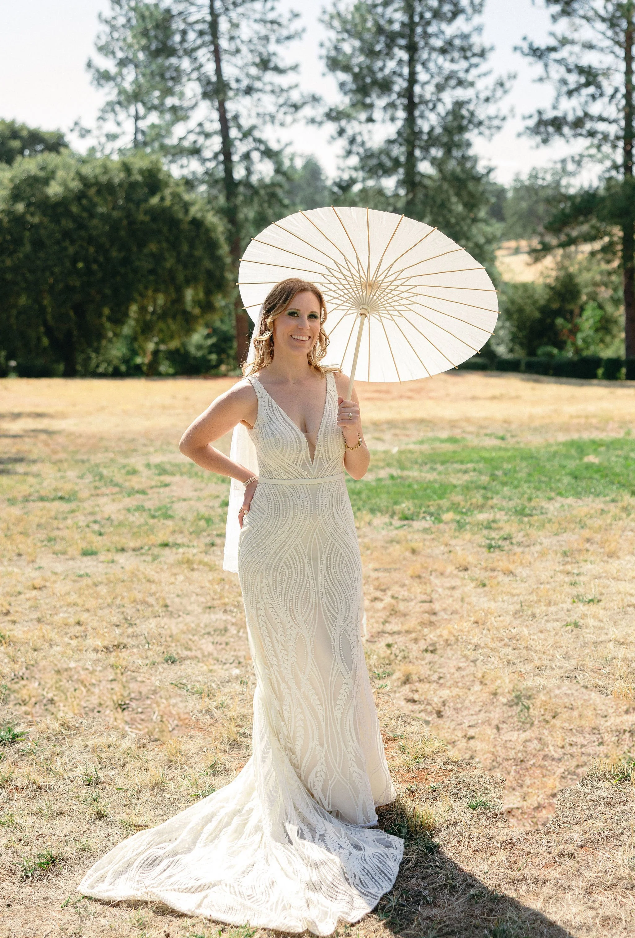 Woman in a white wedding dress holding a parasol outdoors, standing on a grassy area with trees in the background.