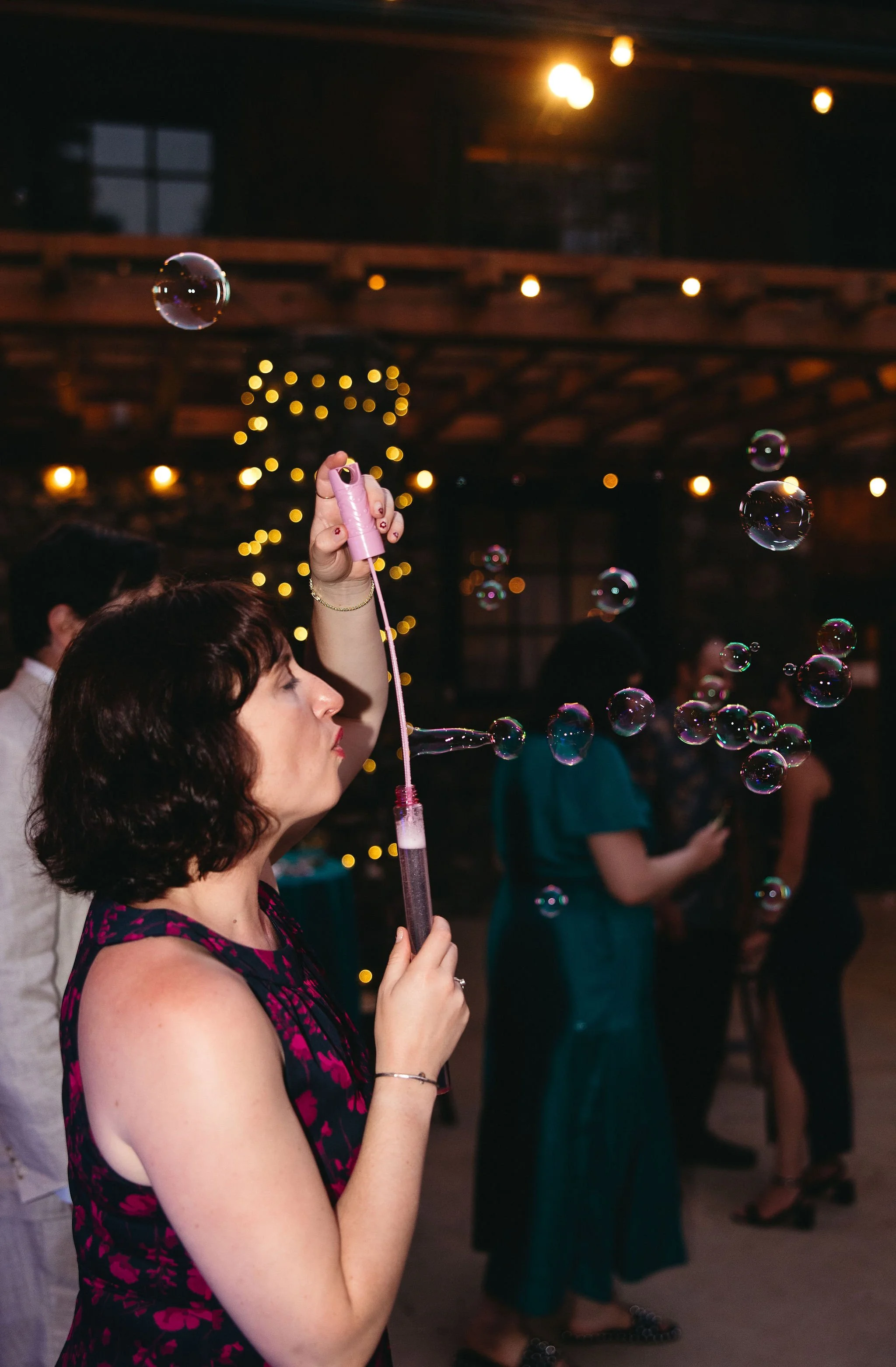 A woman blowing bubbles at a party or celebration in a dimly lit venue with string lights and Christmas tree in the background.