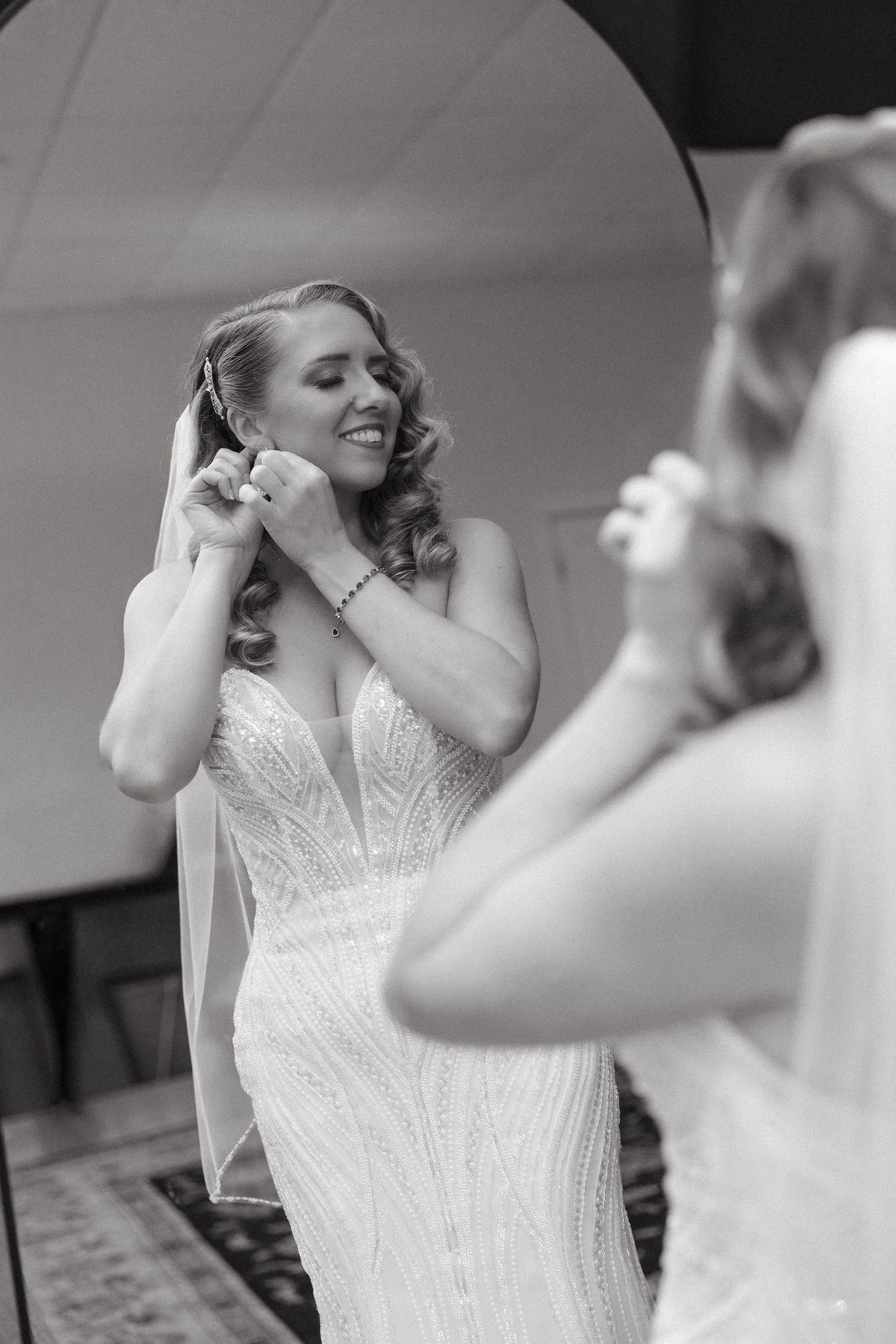 A woman in a wedding dress smiling while putting on earrings, seen in a mirror.