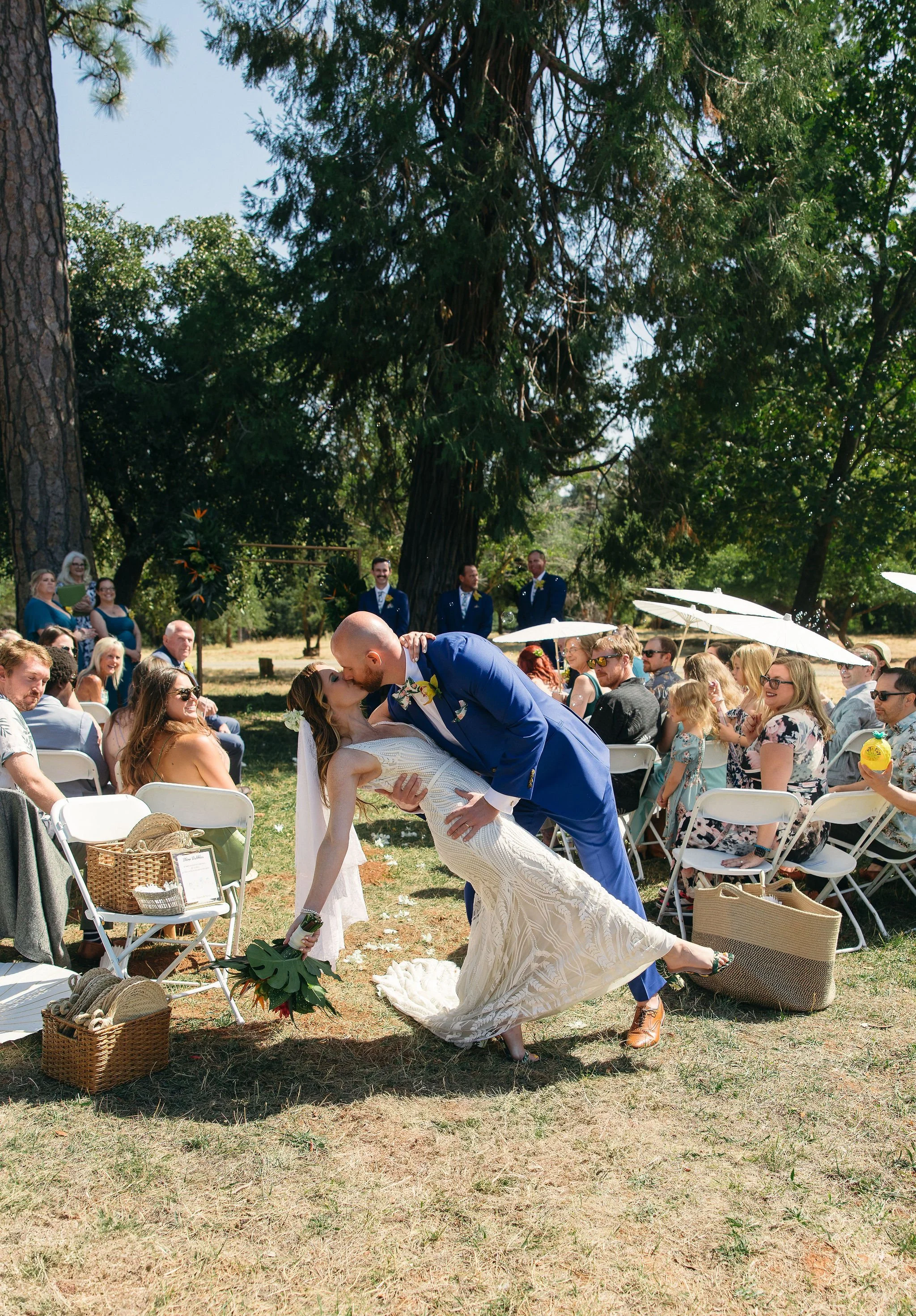 A bride and groom share a kiss during their outdoor wedding ceremony in a park, with guests seated and standing around them, under large trees and sunlight.