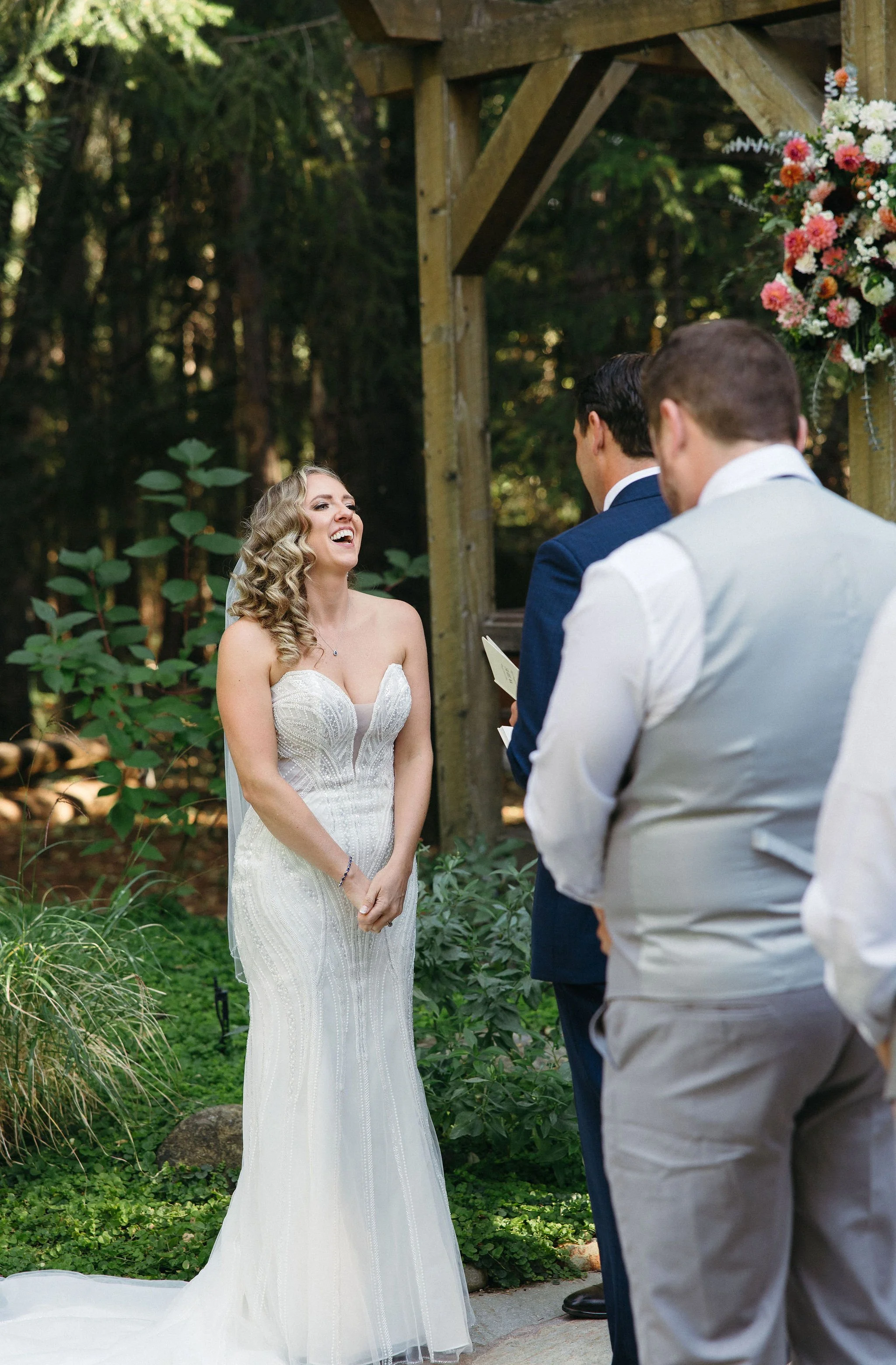 A bride in a white wedding gown during her outdoor wedding ceremony, smiling and laughing, with two groomsmen and a groom standing nearby in a wooded setting.