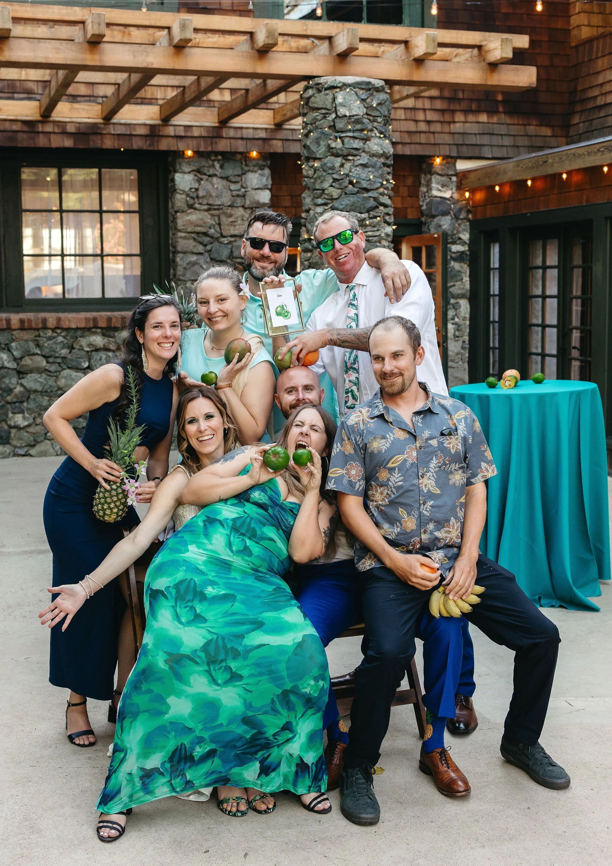 A group of nine people celebrating at a tropical-themed event, wearing colorful clothing with tropical patterns, holding fruits like pineapples and green apples, and posing playfully in an outdoor setting with stone and wood decor.
