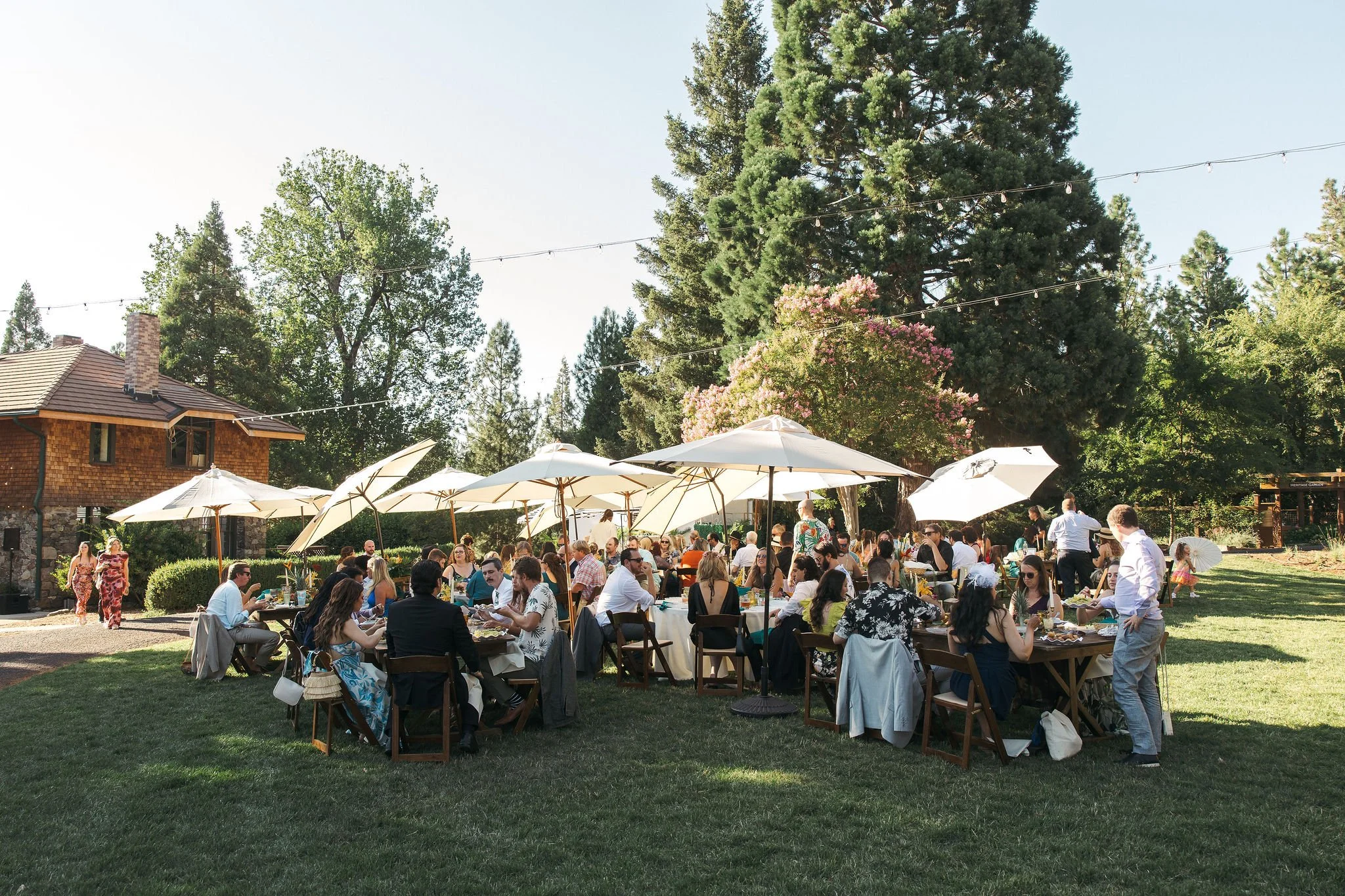 Outdoor garden party with people seated at tables under umbrellas, with trees and a house in the background.