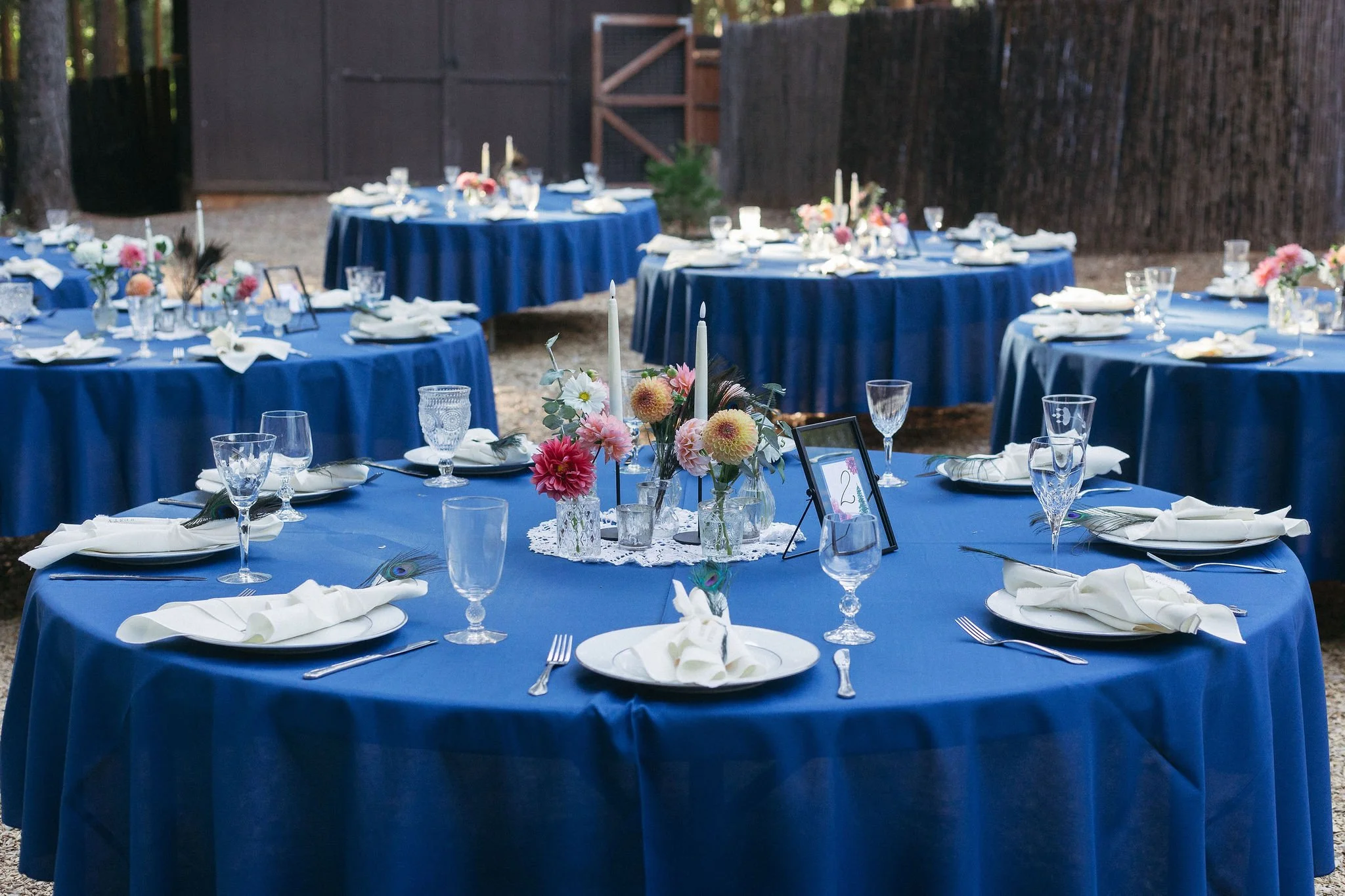 Multiple round tables covered with blue tablecloths, decorated with floral centerpieces, candles, and set with white plates, napkins, and glassware, in an outdoor setting.