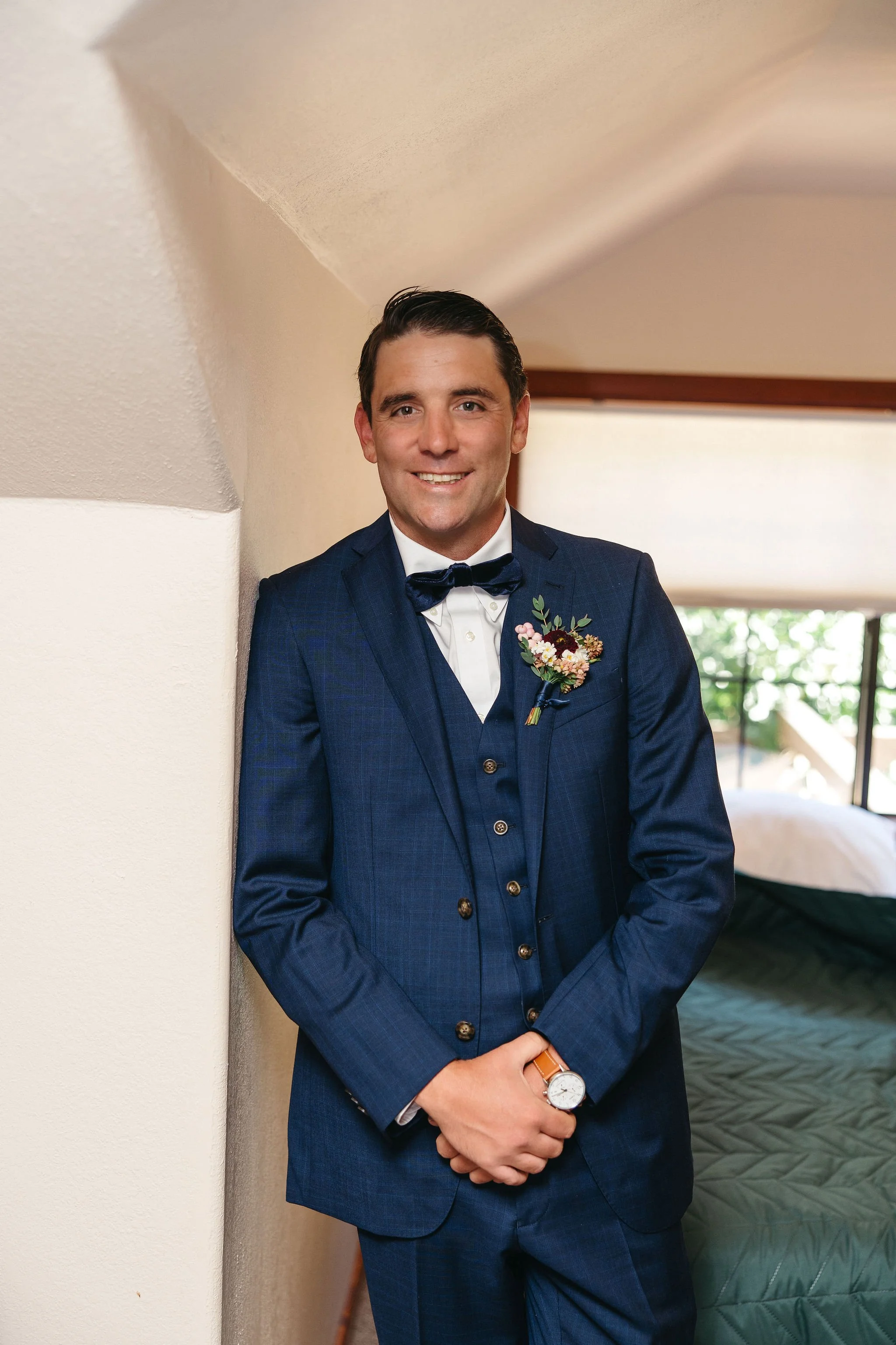 A smiling man in a dark blue suit with a vest, white shirt, and a dark bow tie, standing indoors against a cream-colored wall. He has a boutonniere pinned to his lapel and is wearing a watch with a brown strap.