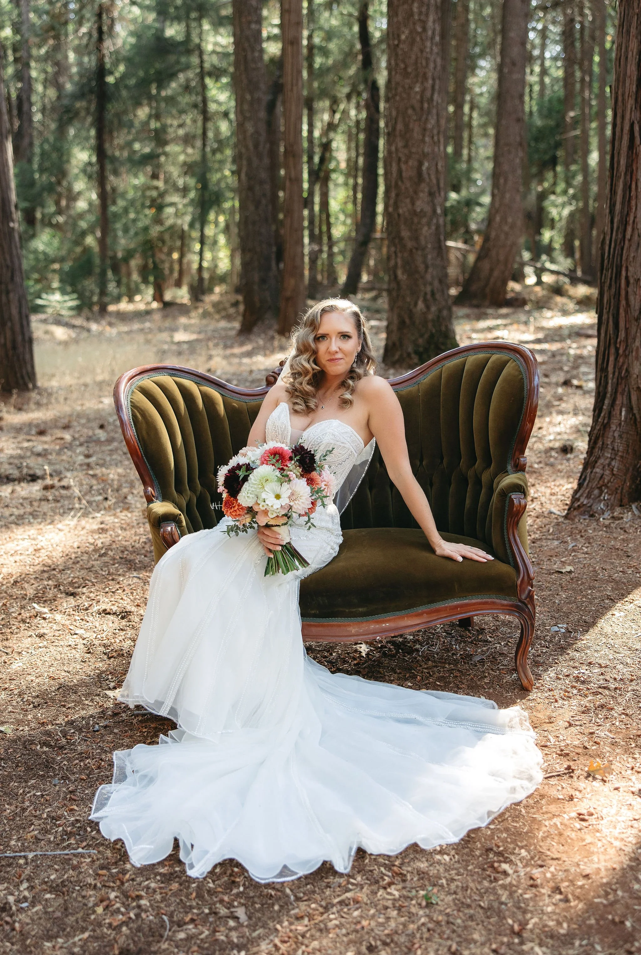 A bride in a white wedding dress sitting on a vintage green velvet settee in a forest, holding a bouquet of flowers, with sunlight filtering through trees.