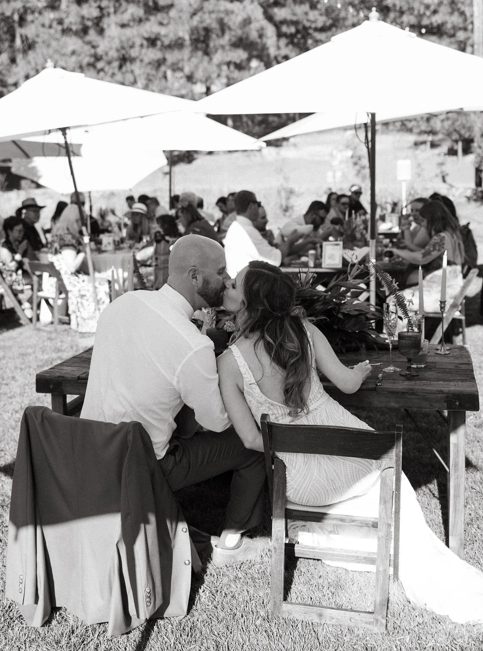 A couple sharing a kiss at their outdoor wedding reception, with guests seated under umbrellas in the background.