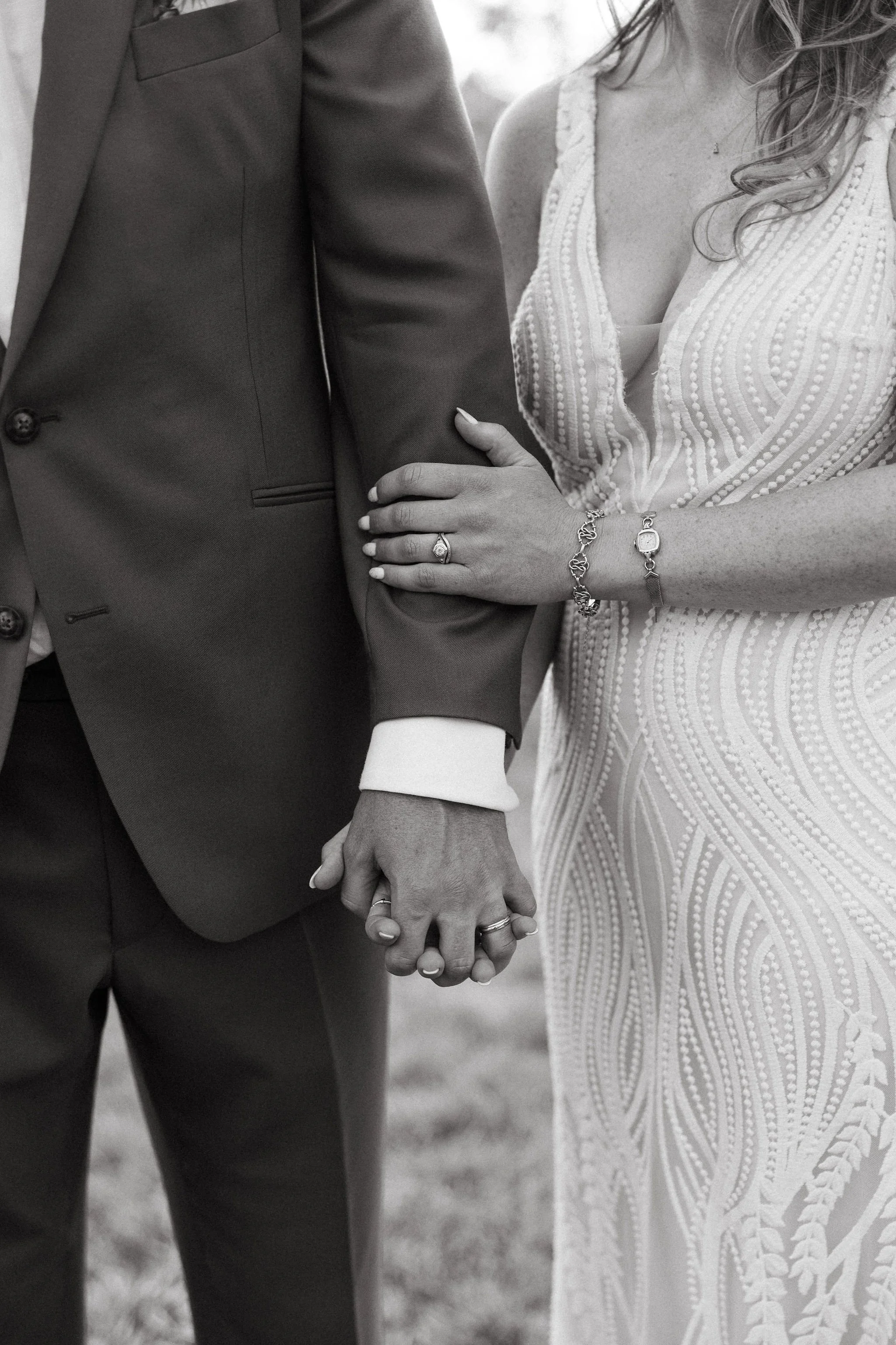 Close-up black and white photo of a couple holding hands, with the woman wearing a white lace dress and jewelry, and the man dressed in a suit.