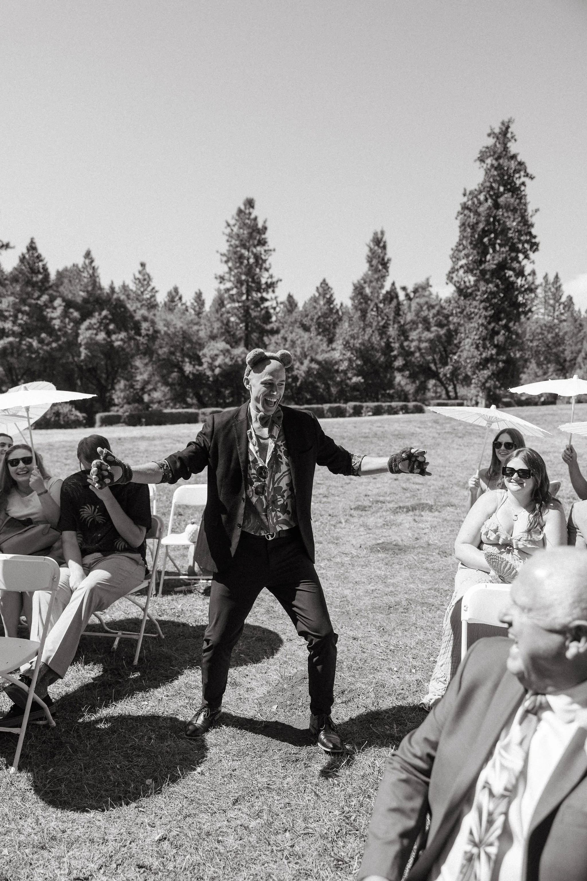A person with animal ears and a patterned shirt performing for an outdoor audience at a garden wedding, with guests sitting on folding chairs and holding umbrellas.