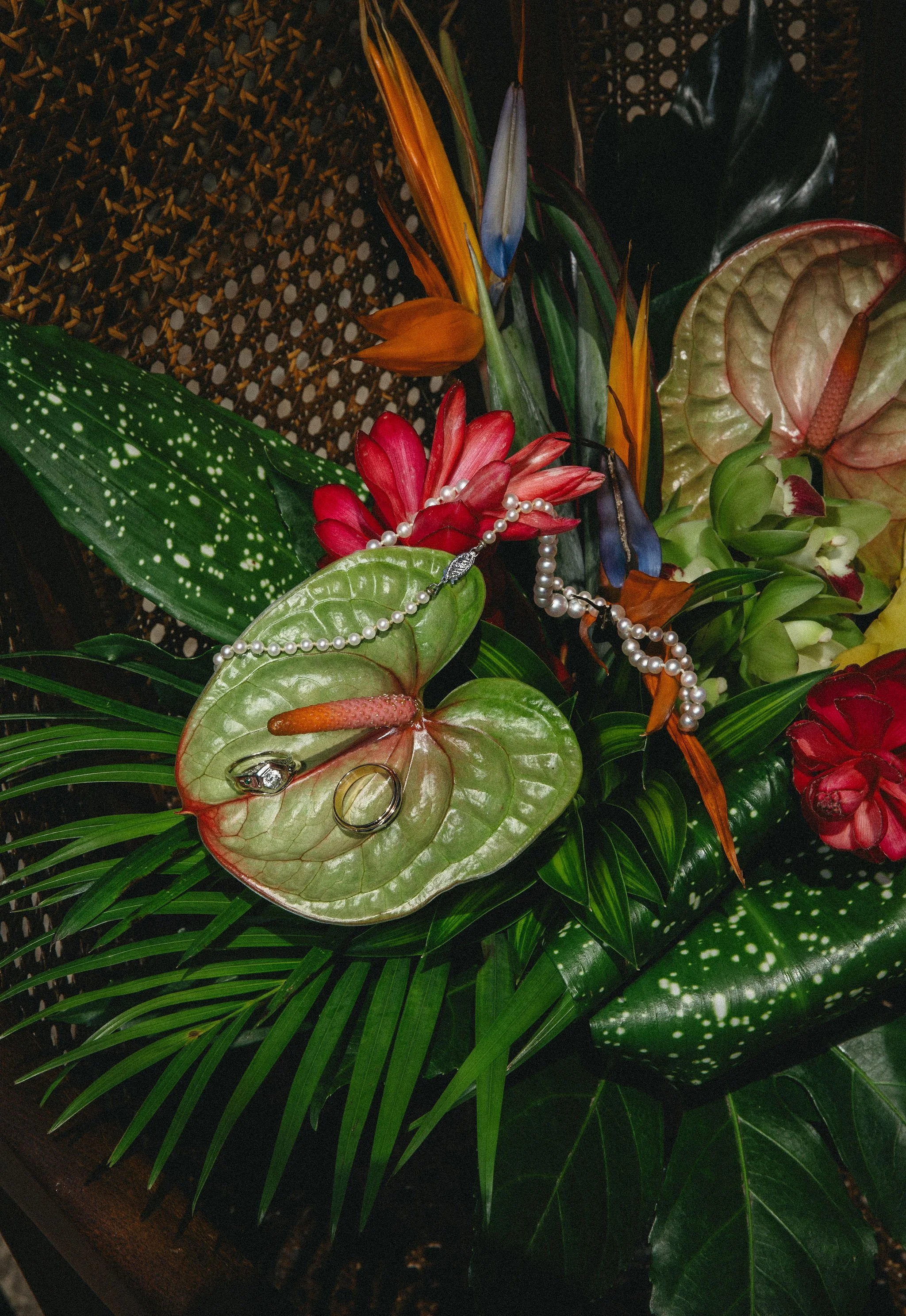 A floral arrangement with various colorful flowers, leaves, and jewelry, including pearl necklaces and rings, on a dark background.