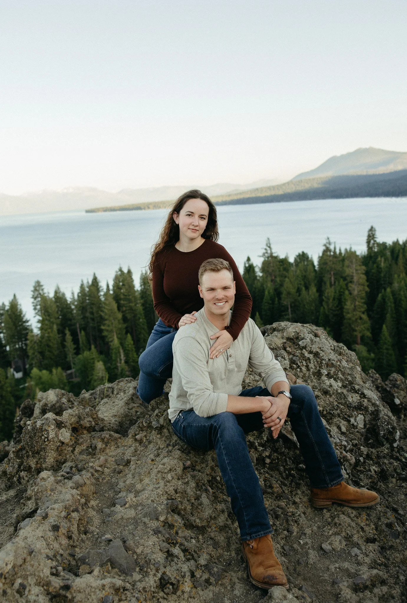 A young man and woman sitting on rocks outdoors with a large lake, forest, and mountains in the background.