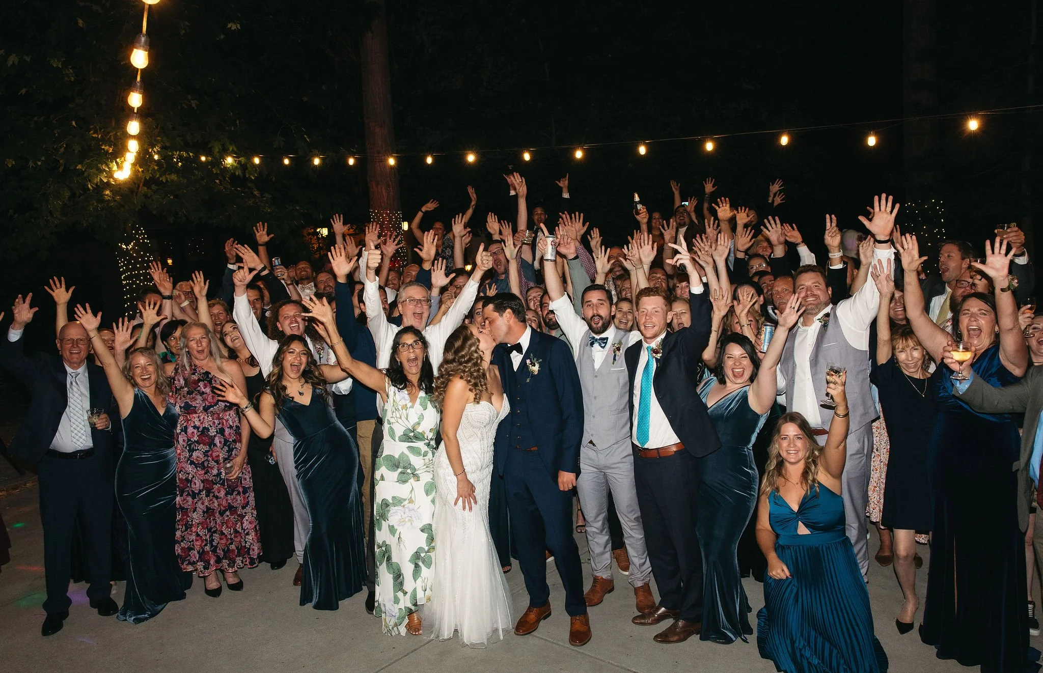 A large group of people celebrating at a wedding reception outdoors at night, with string lights overhead. The bride and groom are in the center, sharing a kiss and surrounded by lively guests raising their hands and holding drinks.