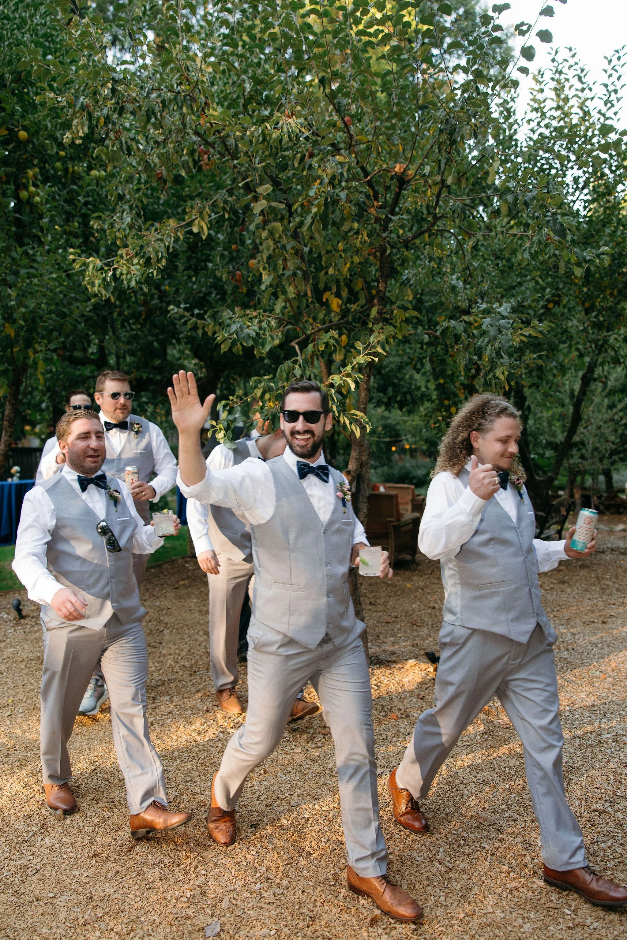 Group of men dressed in coordinated formal outfits, including light gray vests, white shirts, bow ties, and sunglasses, walking outdoors on a gravel path surrounded by greenery, with one man waving and smiling at the camera.