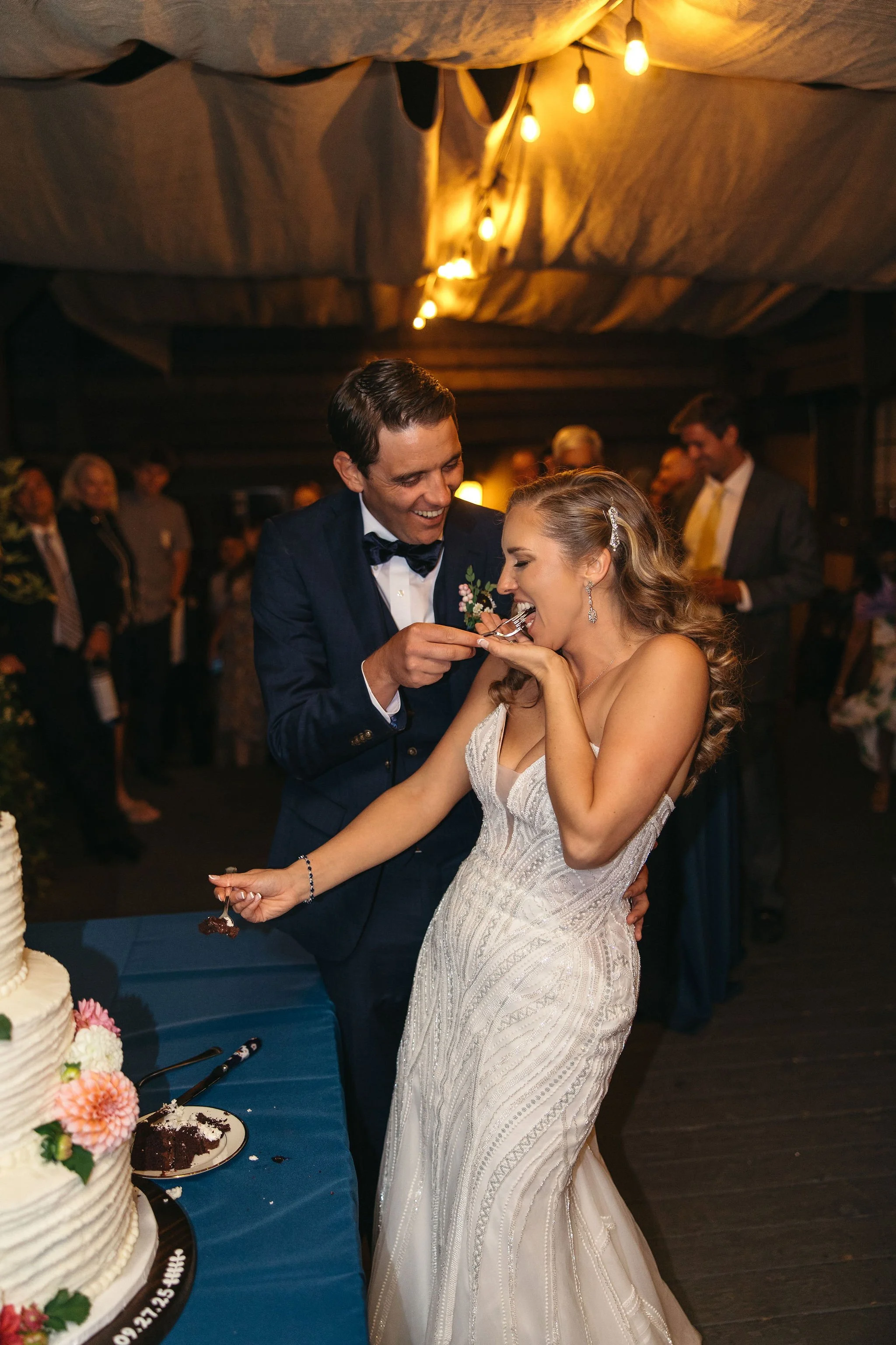 A bride and groom cut a wedding cake together at their wedding reception, with the groom feeding the bride a piece of cake.