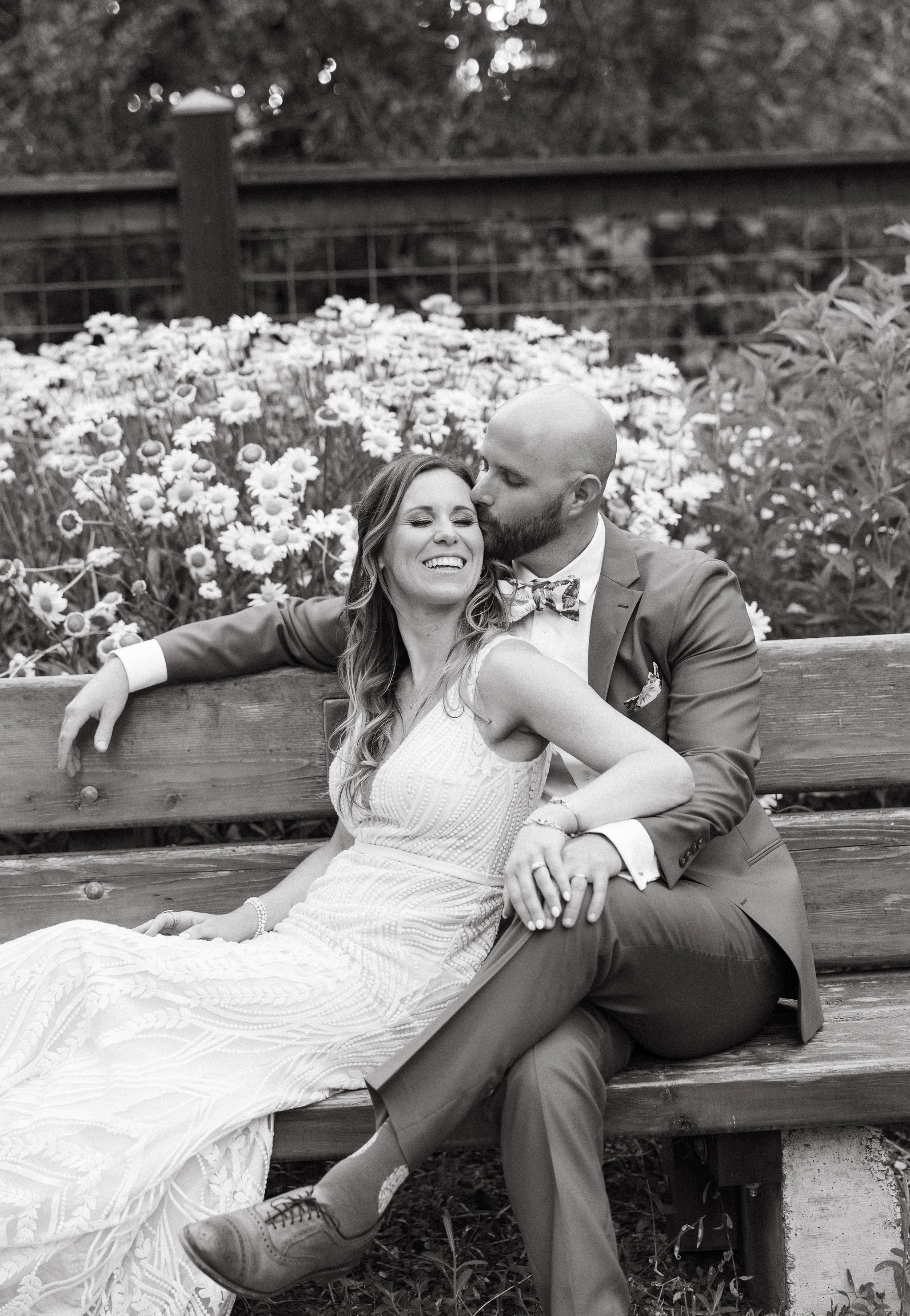 A happy woman and man sitting on a wooden bench in a garden, with the woman smiling and the man kissing her on the cheek, surrounded by flowers.