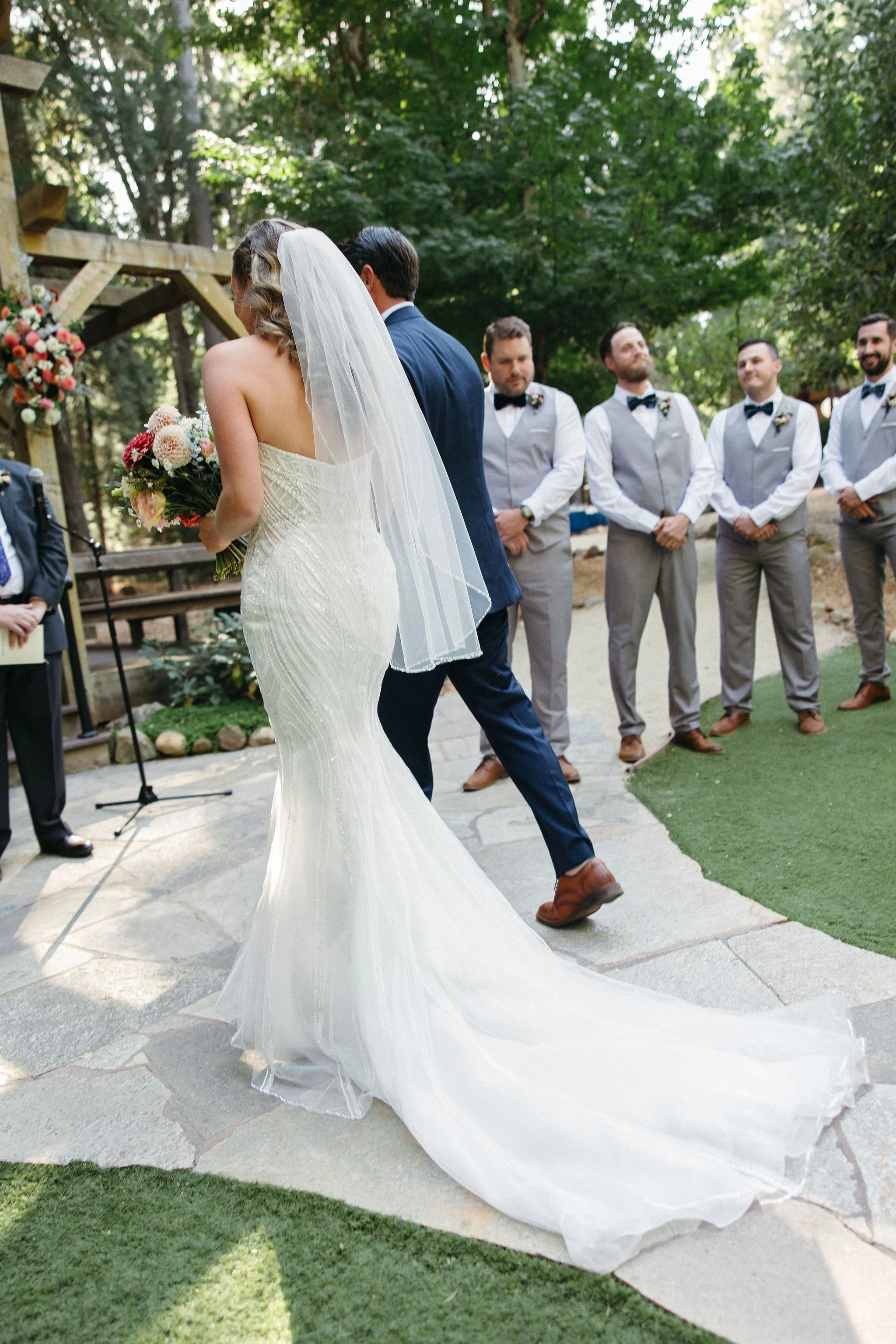 Bride in a white strapless wedding gown with a train, holding a bouquet, and groom in a navy suit walking down the aisle at an outdoor wedding ceremony surrounded by groomsmen in gray vests and black bow ties, with a wooded backdrop.