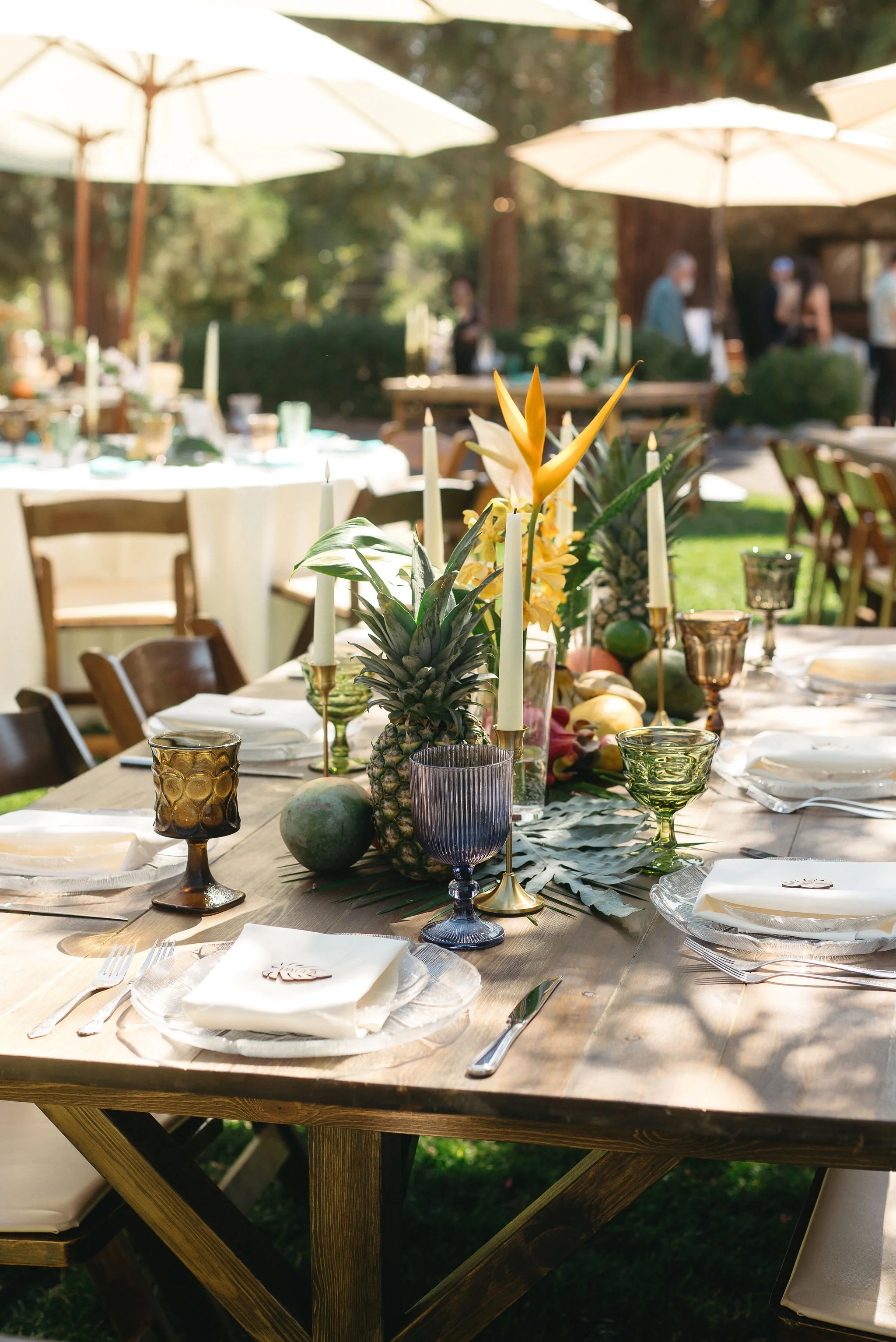 Outdoor table set for a celebration with fruit, candles, and decorative glassware during daytime.