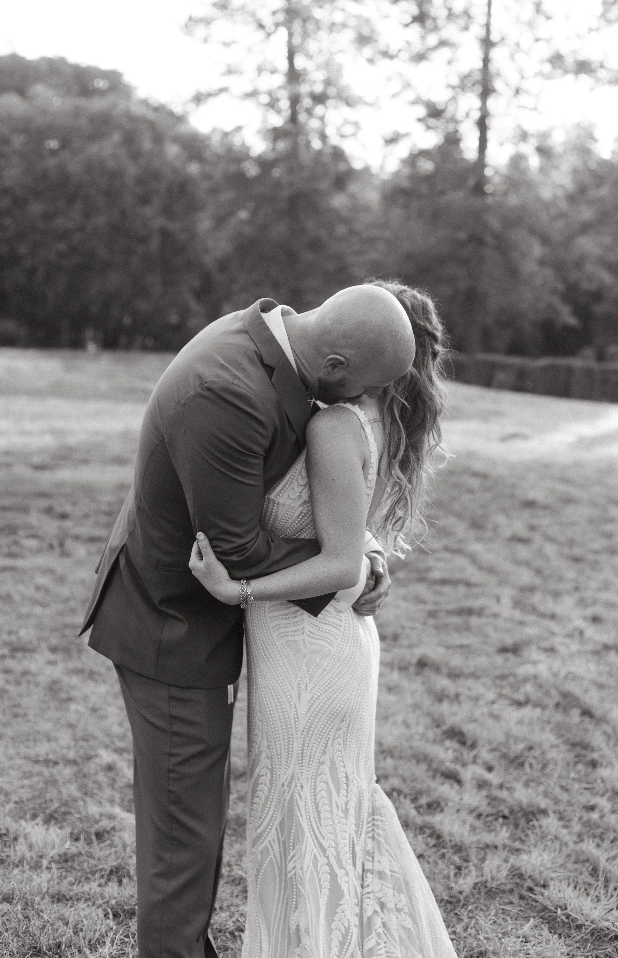 A black-and-white photo of a couple kissing outdoors on a grassy field. The man is wearing a suit, and the woman is wearing a white lace wedding dress with her long, wavy hair loose. The background shows trees and a slightly blurred landscape.