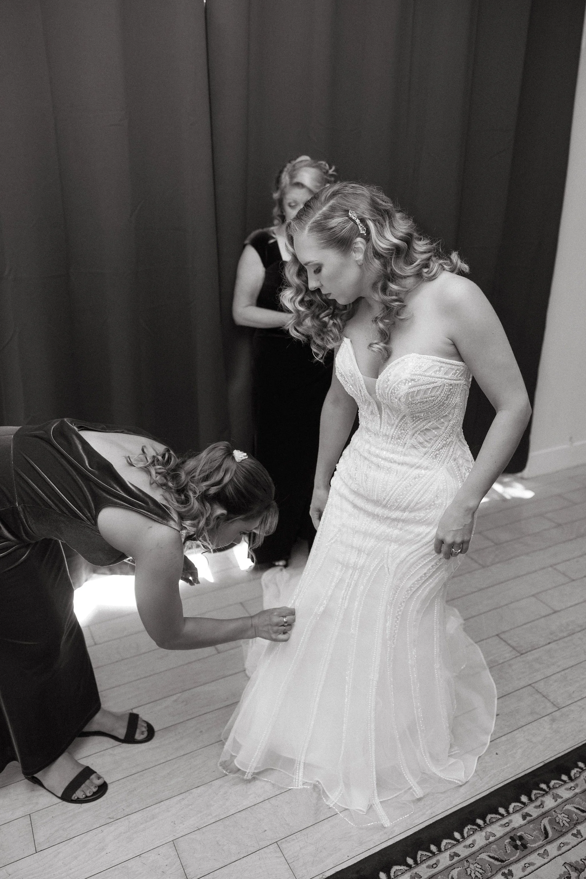 A woman in a wedding dress having her shoe adjusted by a helper, with another woman in the background.