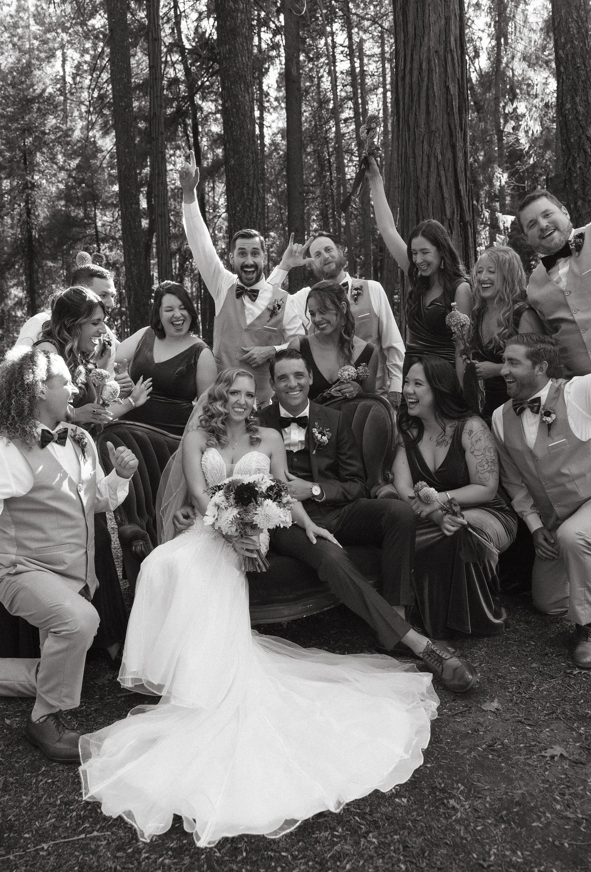 A wedding celebration in a forest with a group of people, including the bride and groom sitting on a chair in the center, surrounded by friends who are cheering and smiling.
