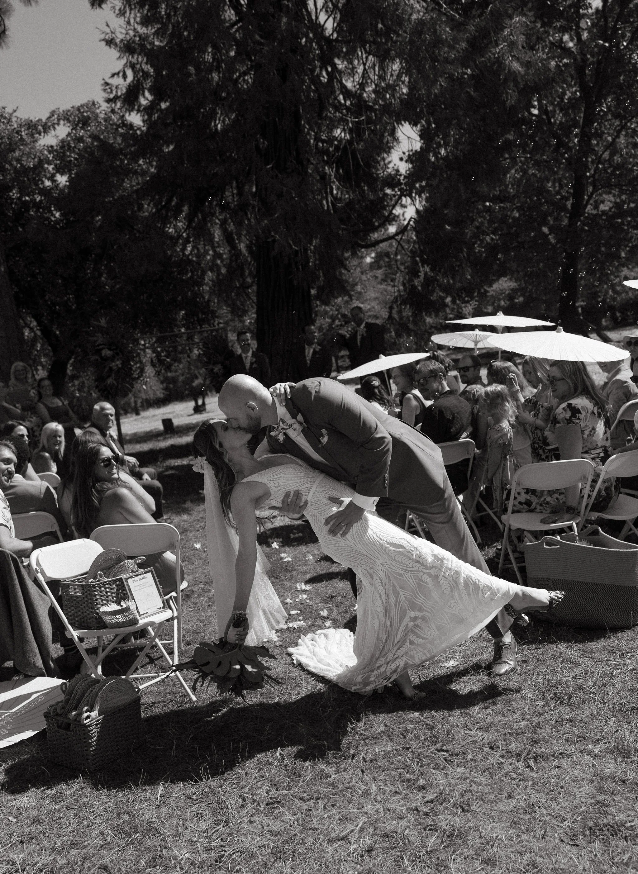 A bride and groom sharing a kiss during an outdoor wedding ceremony. The groom is dipping the bride, who is holding a bouquet, while guests watch and sit in chairs under umbrellas in a shaded area with trees.