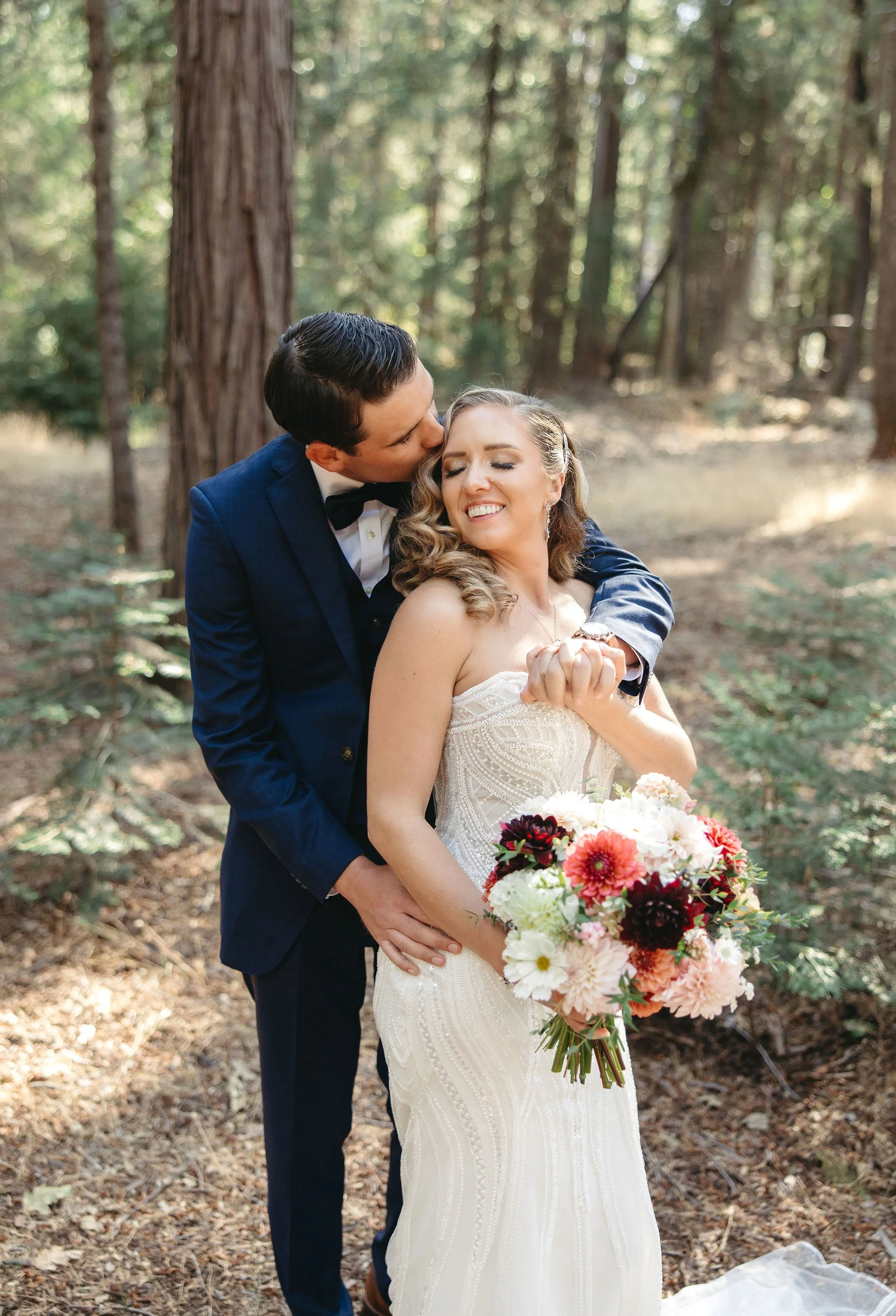 A bride in a white wedding gown holding a bouquet of flowers, being embraced and kissed on the cheek by a groom in a dark suit and bow tie in a forest setting.