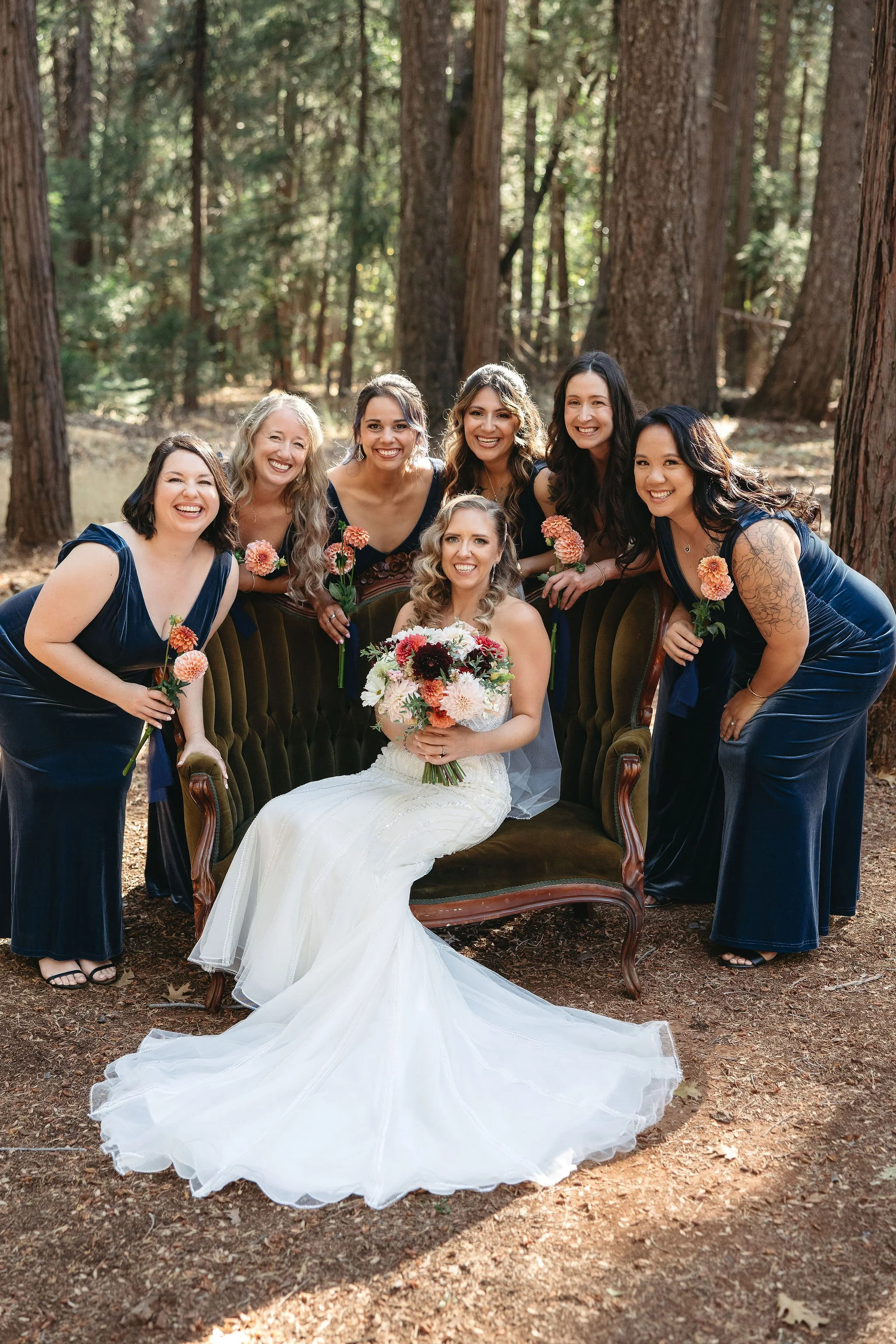 A bride in a white wedding dress sitting on a vintage sofa in a forest, surrounded by her bridesmaids in navy blue dresses, all smiling and holding pink flowers.