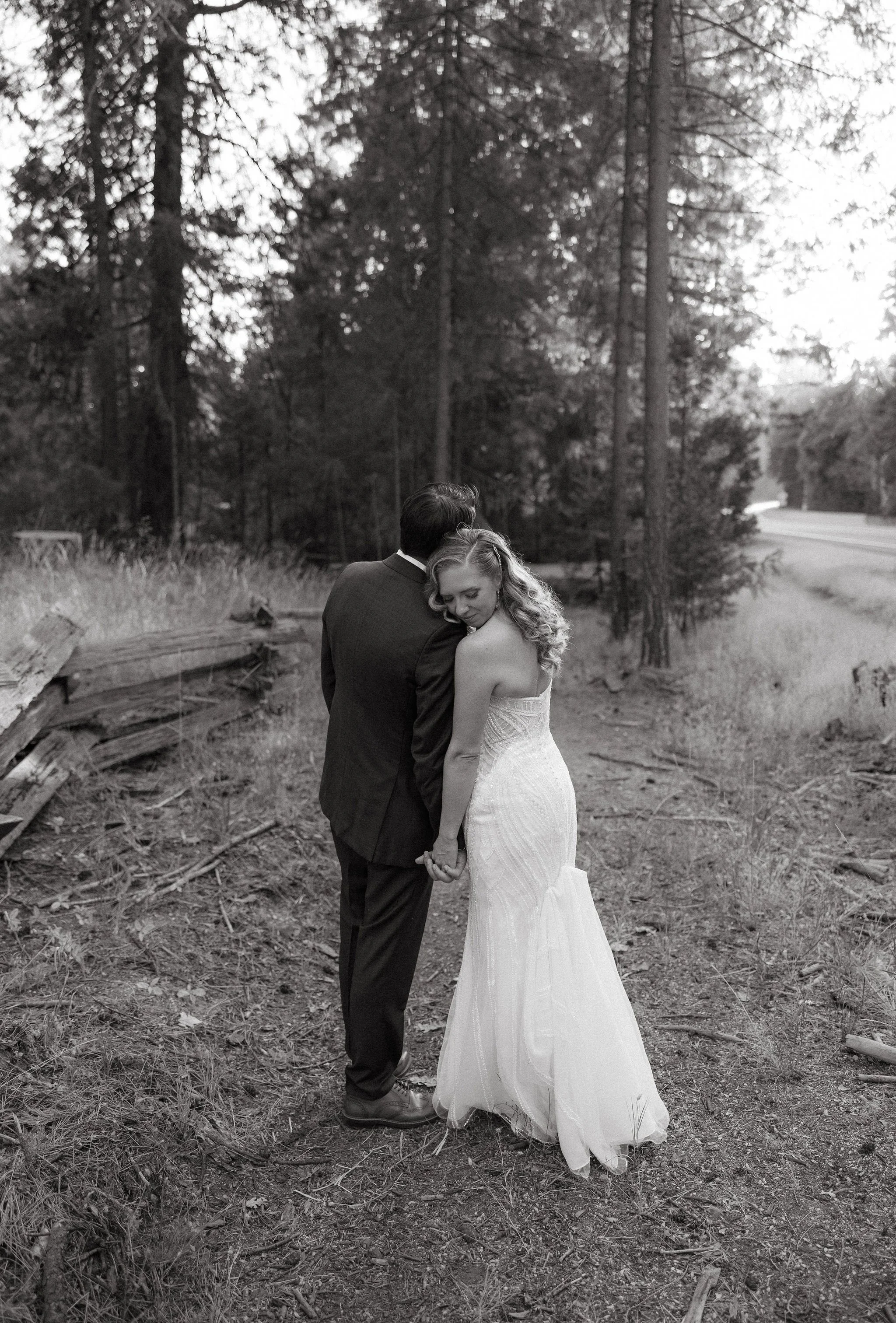 A black and white photo of a bride and groom standing together outdoors in a wooded area, holding hands and leaning into each other.