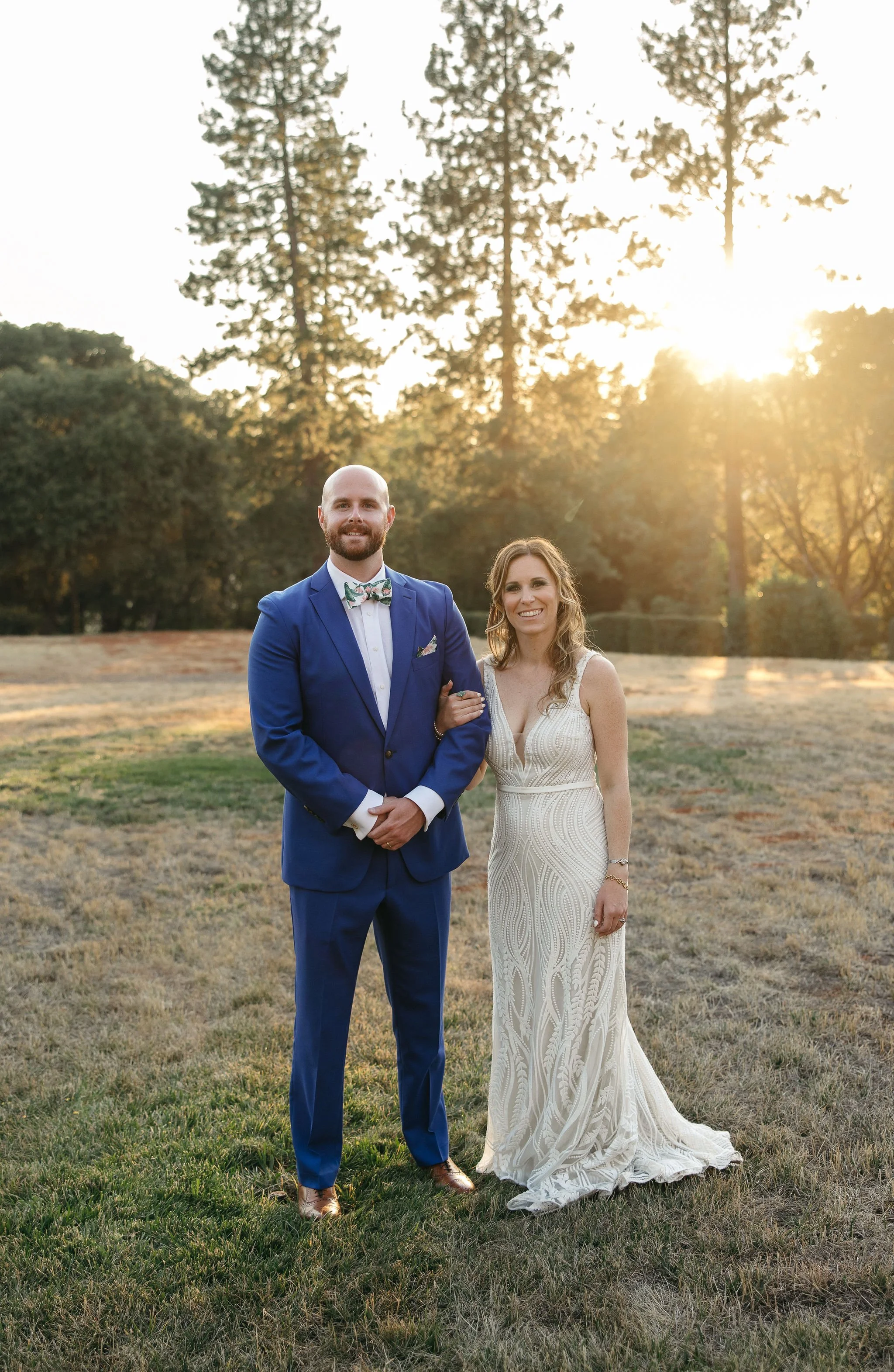 A newlywed couple standing outdoors during sunset, dressed in formal wedding attire. The groom is wearing a blue suit with a bow tie, and the bride is in a white, sleeveless wedding gown. They are smiling and holding hands, with trees and the setting