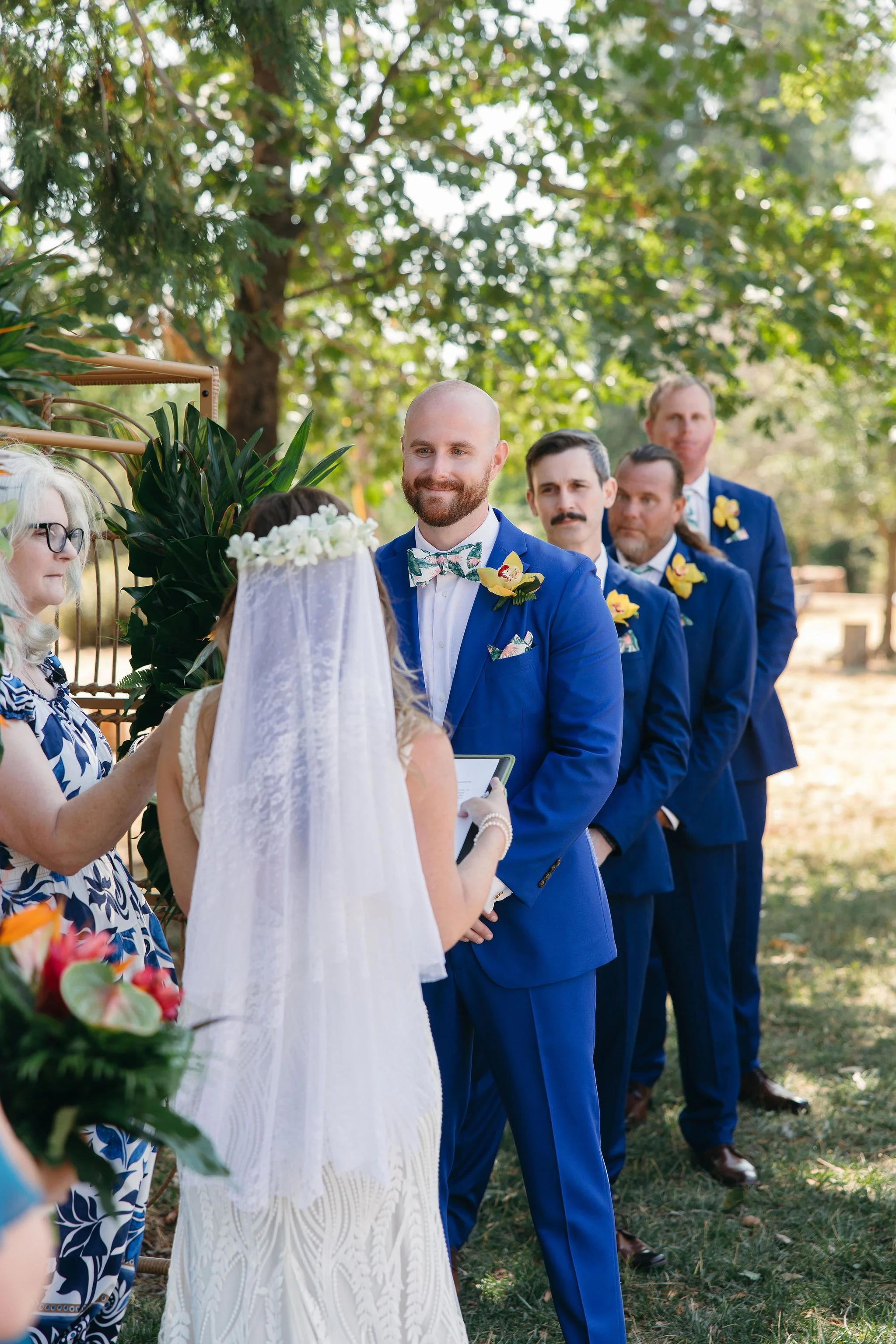 A wedding ceremony outdoors with a bride and groom facing each other, surrounded by bridesmaids and groomsmen in blue suits, under trees with sunshine filtering through.