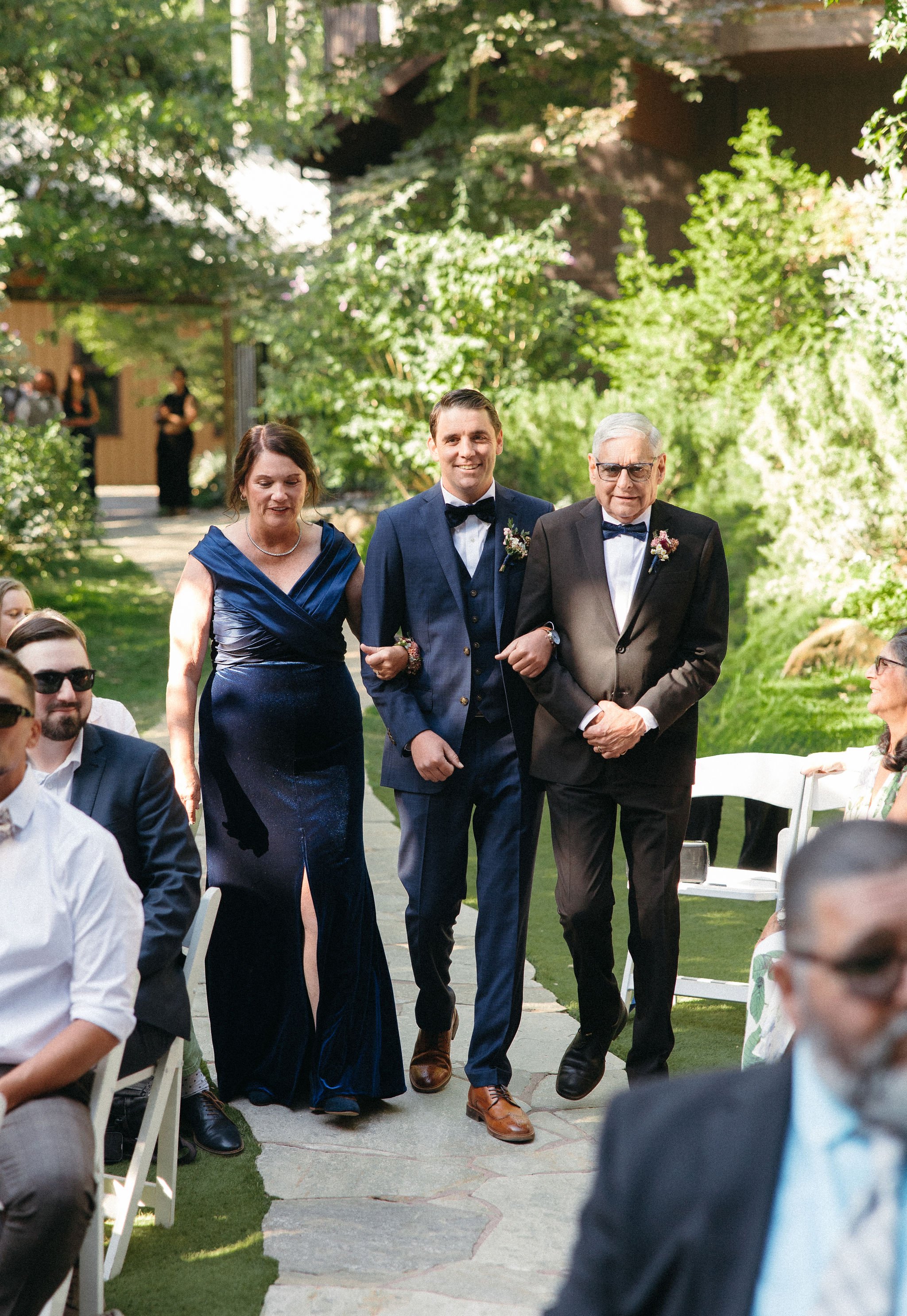 A man in a blue suit and bow tie walks down the aisle with two older men, one in a black tuxedo and the other in a navy vest and pants, at an outdoor wedding ceremony. A woman in a dark blue dress walks beside him. Guests seated on either side watch 