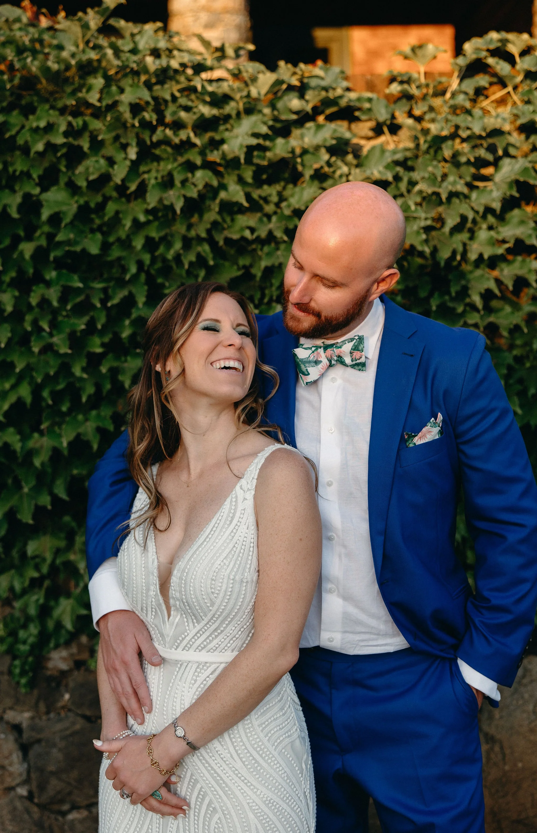 A smiling woman in a white wedding dress and a man in a blue suit with a floral bow tie sharing a joyful moment outdoors, standing in front of a bush with green leaves.