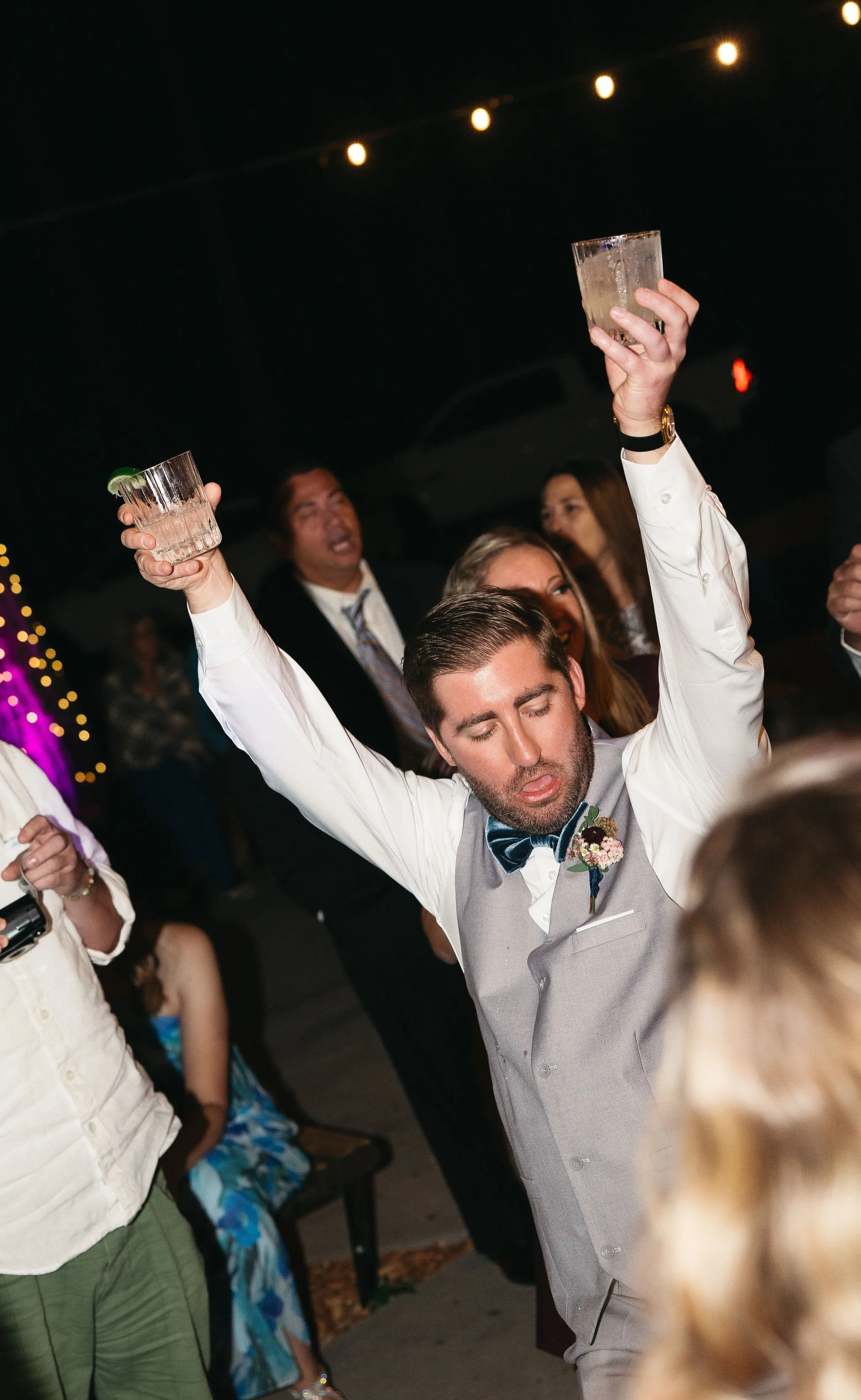 A groom with closed eyes and a light gray vest, white shirt, black bow tie, and boutonniere, raising his glass while dancing at a lively celebration with other guests in the background.