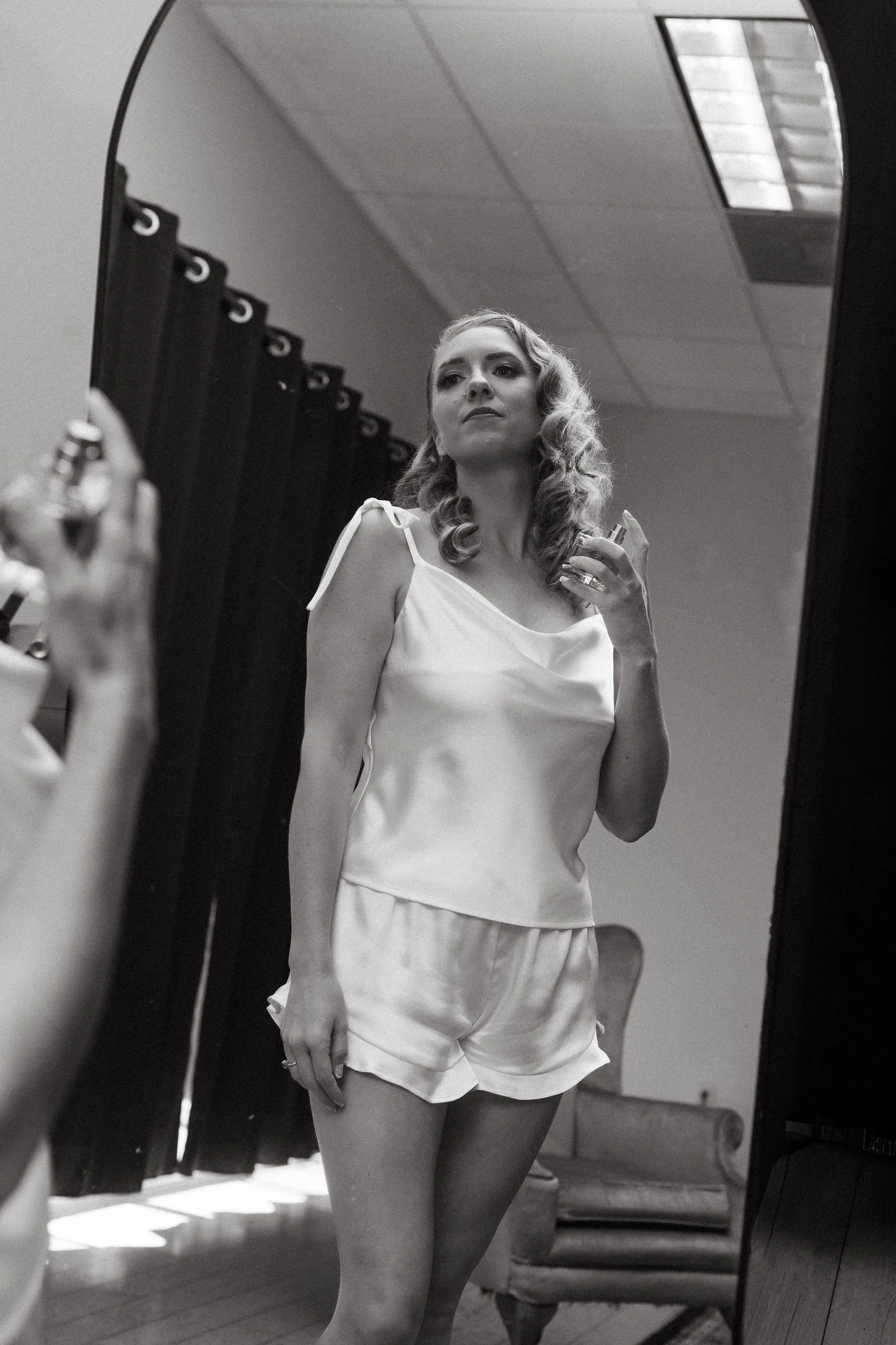 A woman with curly hair is looking at herself in a mirror, holding a perfume bottle in her right hand, wearing a satin camisole and shorts, in a room with curtains and a vintage chair.