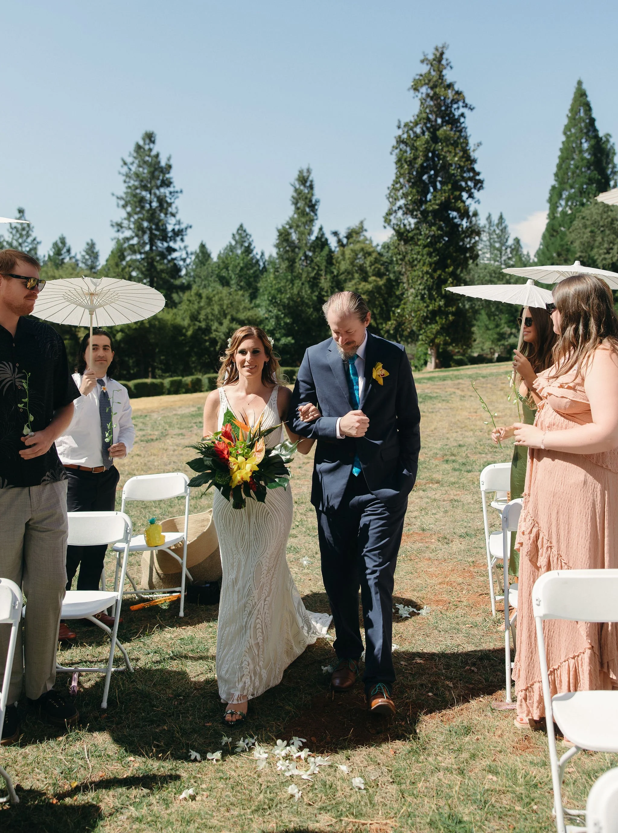 A couple walking down the aisle at an outdoor wedding ceremony, with the bride holding a bouquet and the groom with his arm linked with hers, surrounded by guests holding umbrellas on a sunny day.