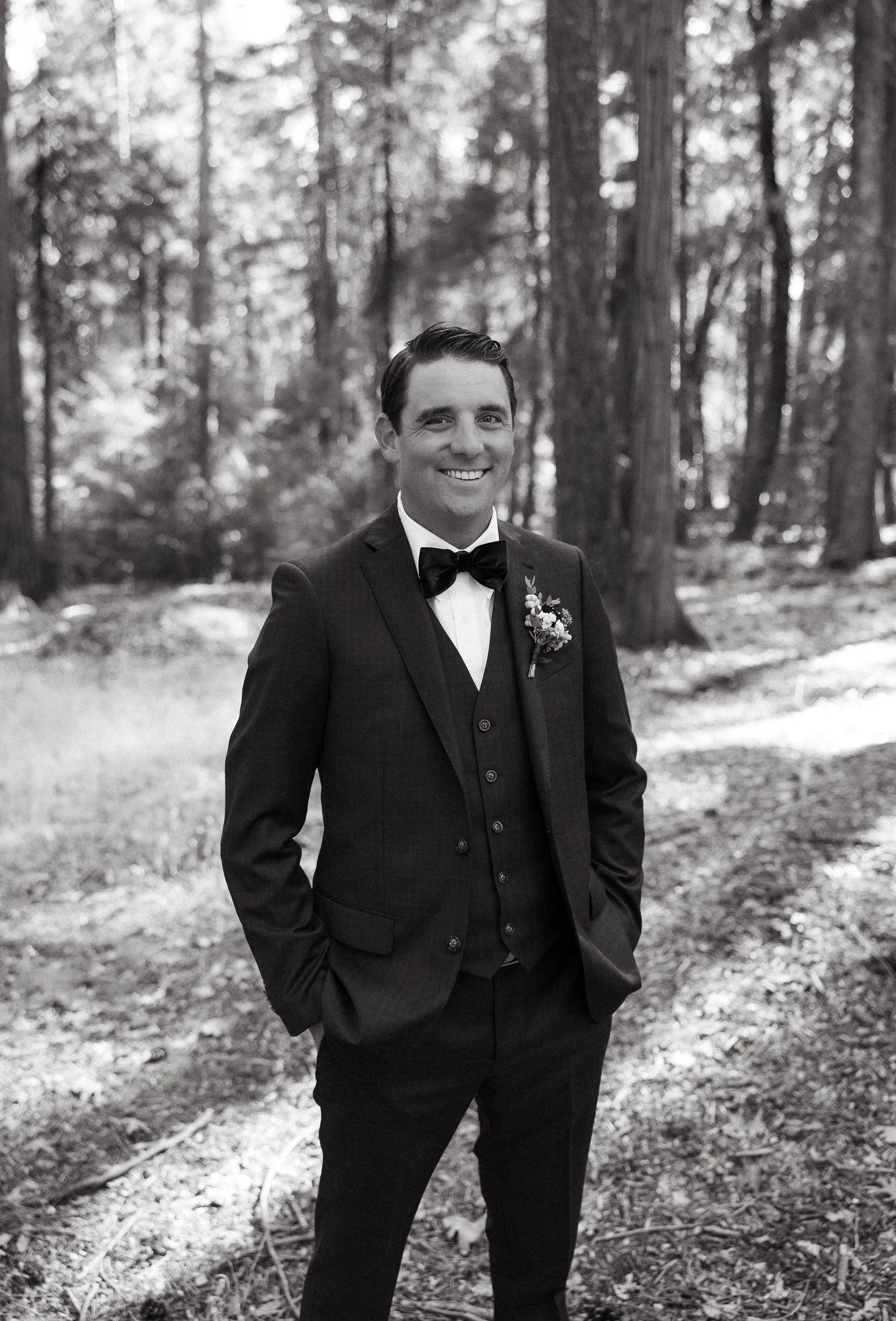 A man in a tuxedo with a bow tie and boutonniere, standing outdoors in a forest with tall trees, smiling at the camera.