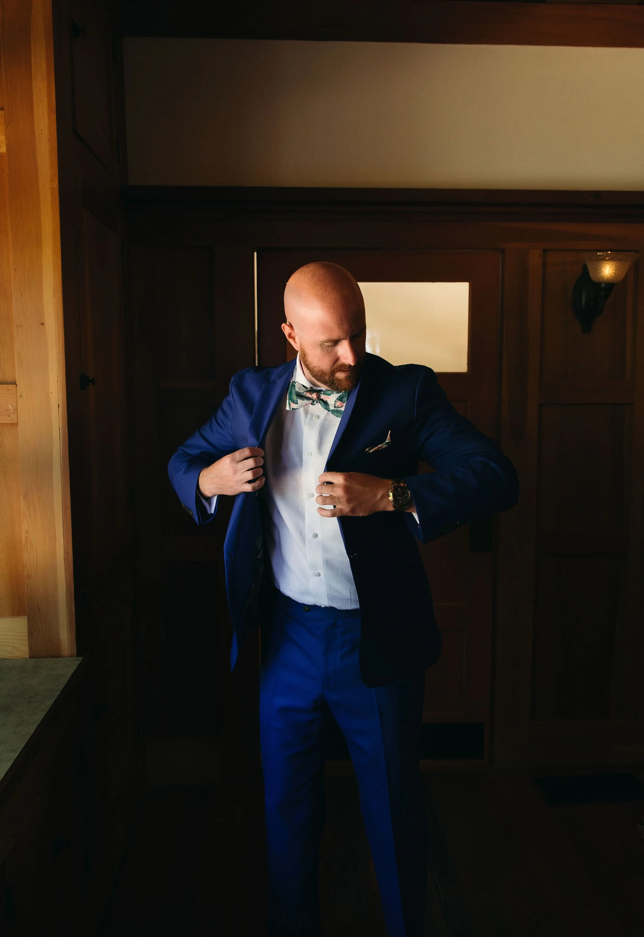 A man adjusting his blue suit jacket indoors, wearing a white shirt, patterned bow tie, and a watch, with wooden walls and a small window in the background.