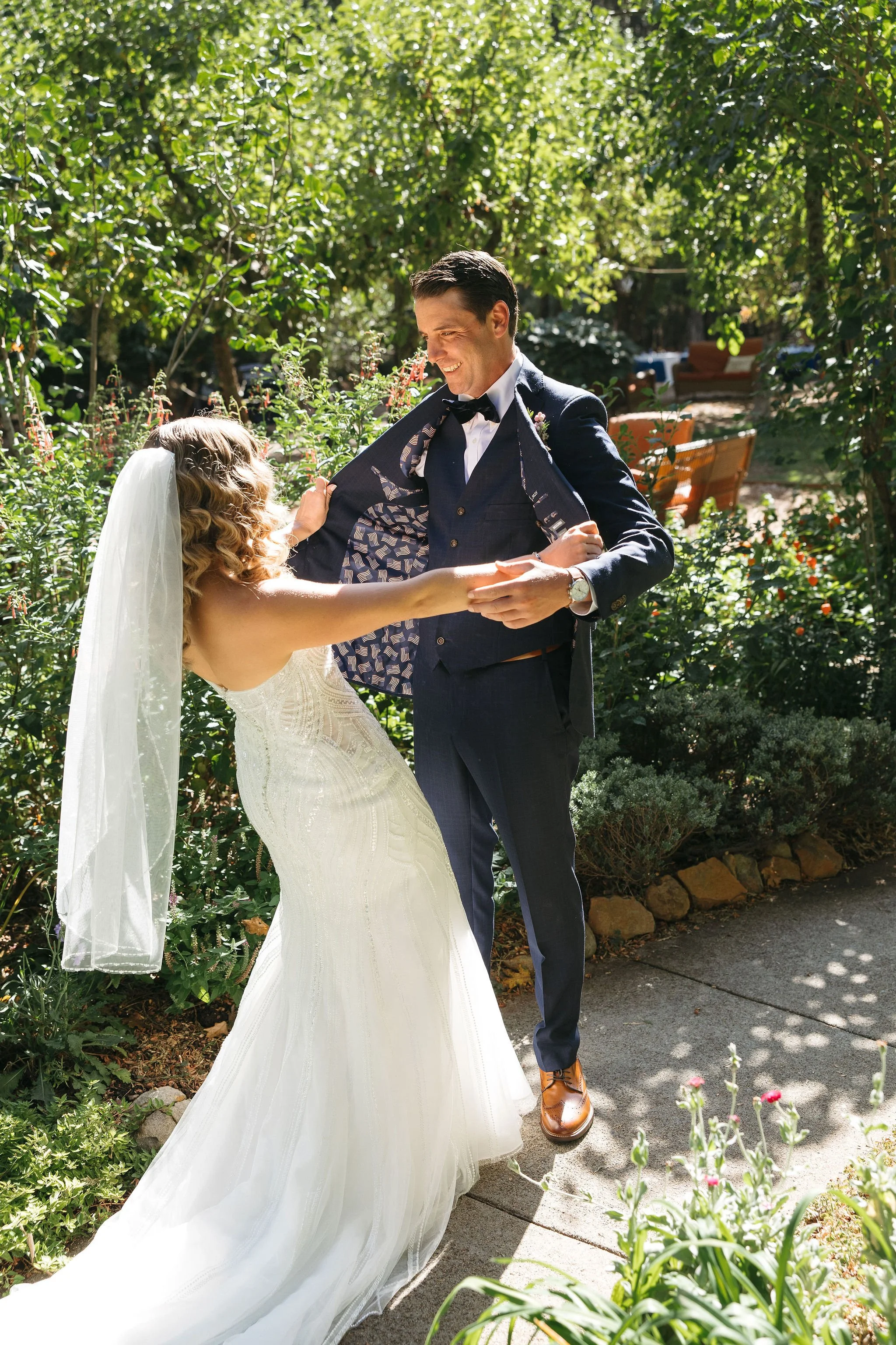 A bride and groom dancing outdoors in a lush garden, with the bride in a white wedding gown and veil, and the groom in a dark suit with a bow tie, smiling and holding hands.