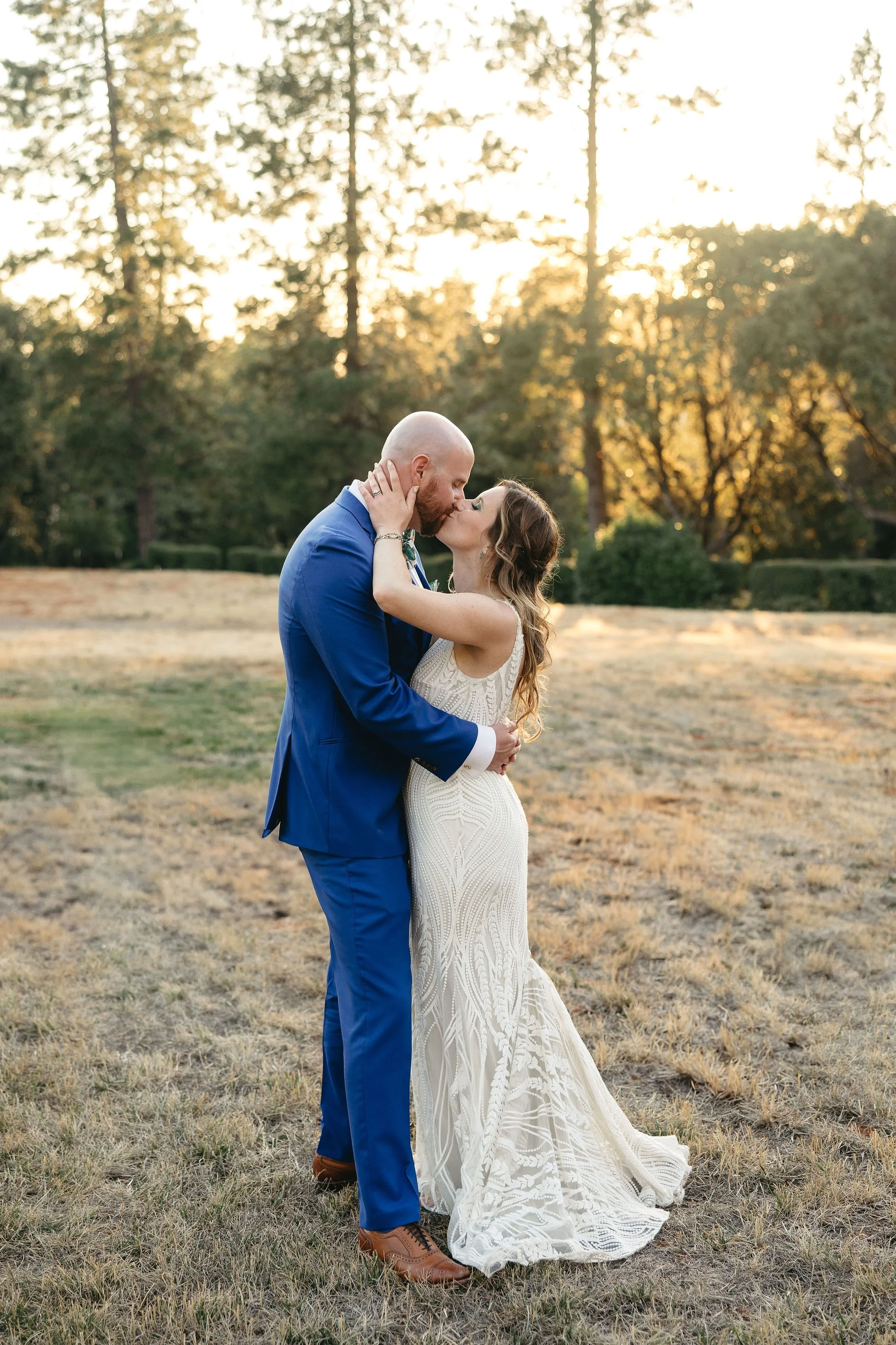 A wedding couple kissing outdoors at sunset, with trees in the background, the bride in a white gown and the groom in a blue suit.