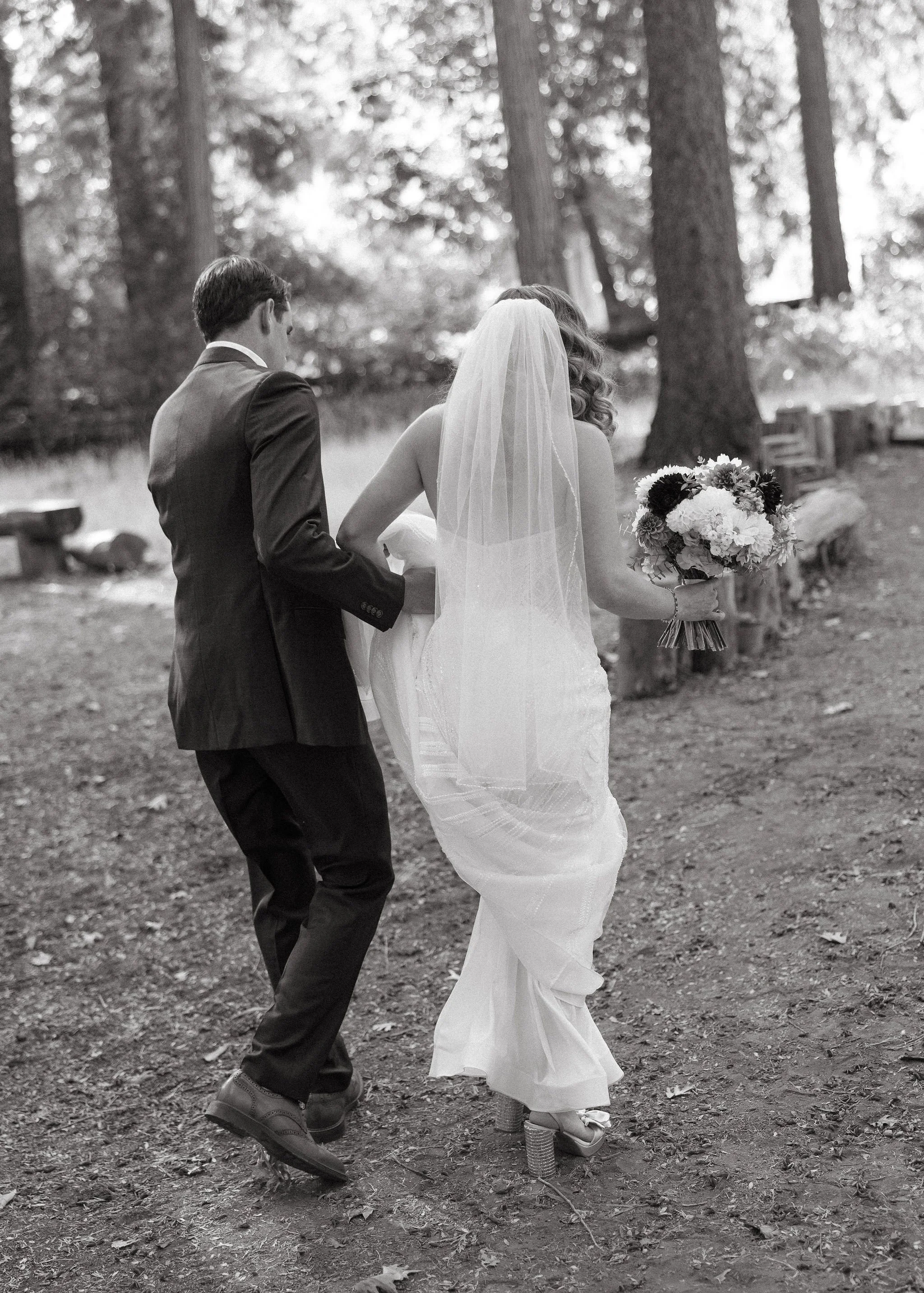Black and white photo of a bride and groom walking in a wooded outdoor area. The bride is wearing a wedding dress and veil, holding a bouquet of flowers, and standing on high heels. The groom is in a suit and dress shoes, holding the bride's dress as