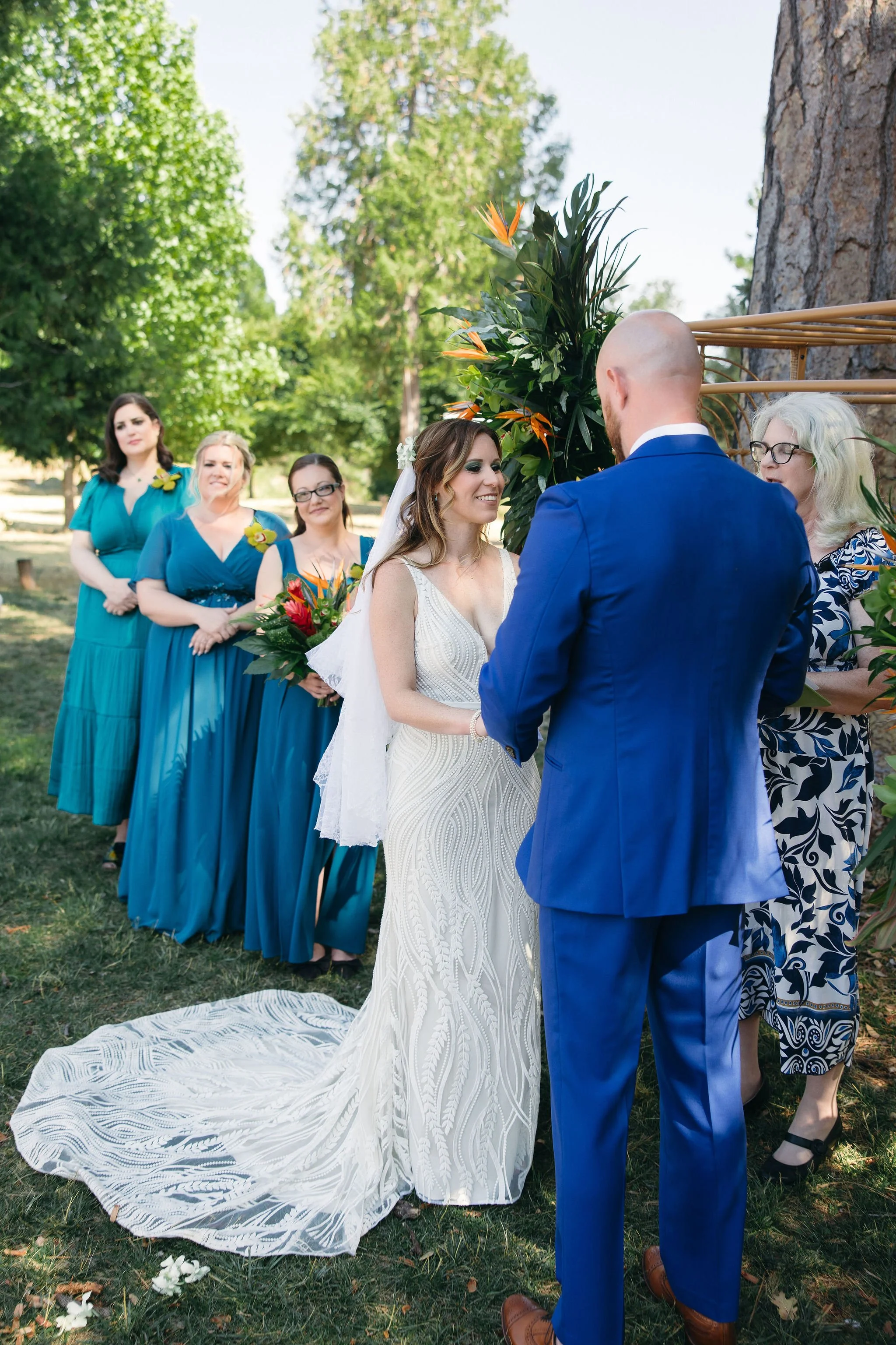 A wedding ceremony outdoors with the bride and groom exchanging vows. The bride wears a white wedding dress with a train and veil, holding a bouquet of orange and green flowers, while the groom is dressed in a blue suit. Bridesmaids in teal dresses s