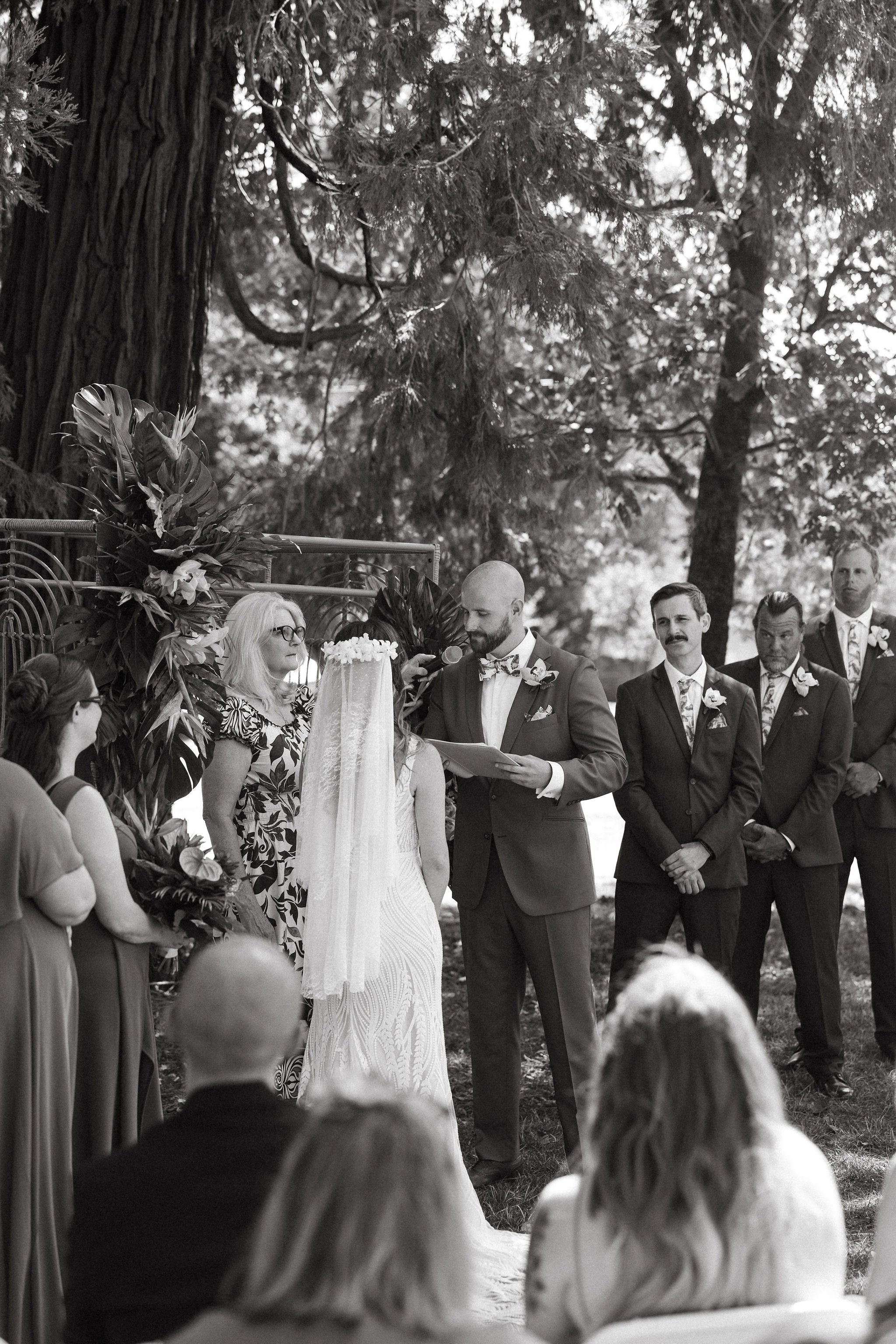 A black and white photo of a wedding ceremony outdoors beneath trees, with a couple exchanging vows and a group of groomsmen and guests watching.